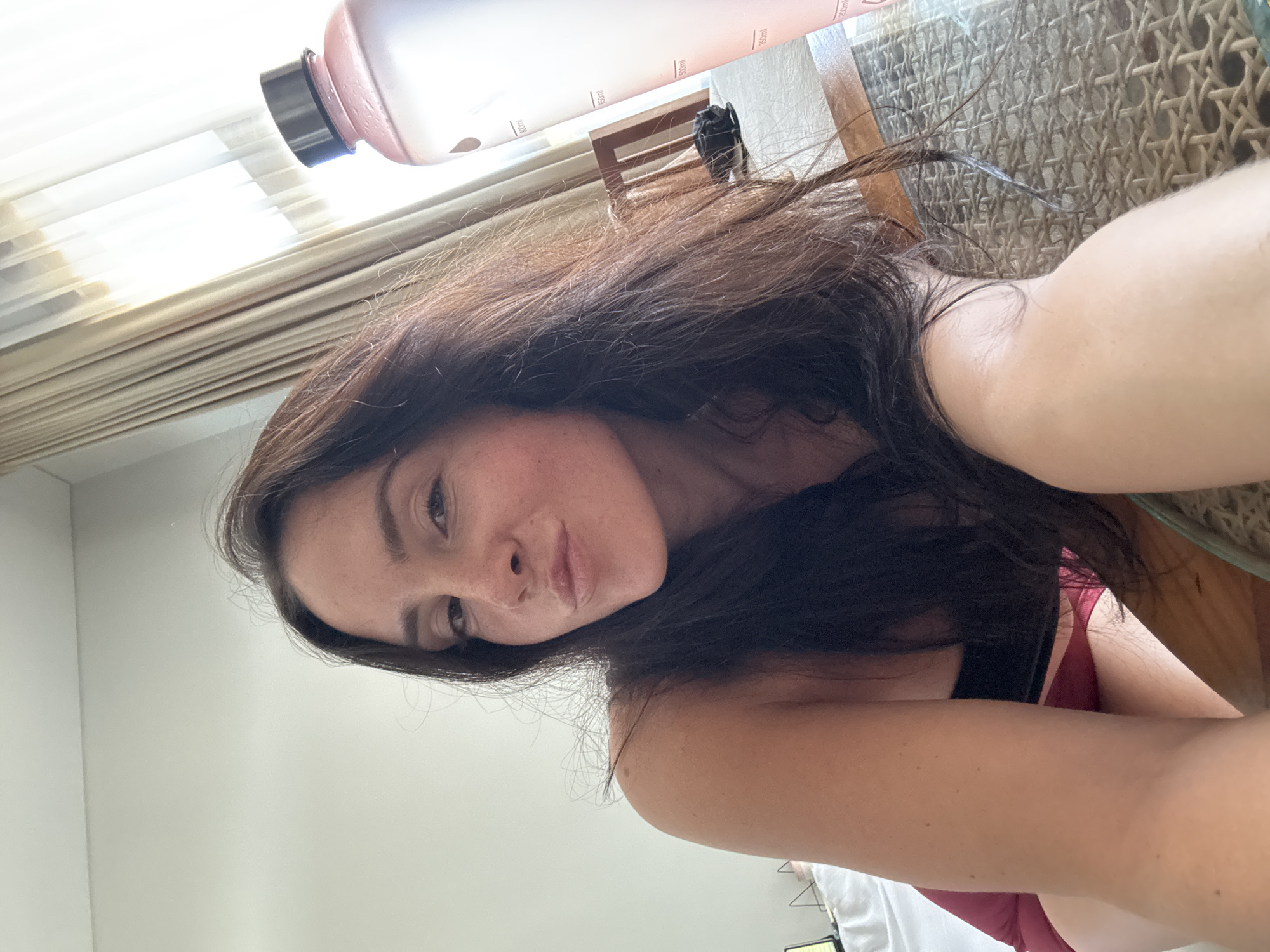 A woman is lying on a table in a room. She has long, dark hair and is wearing a dark top and red shorts. She is looking at the camera and making a pouty face. There is a large water bottle to her right. The room has curtains in the background.