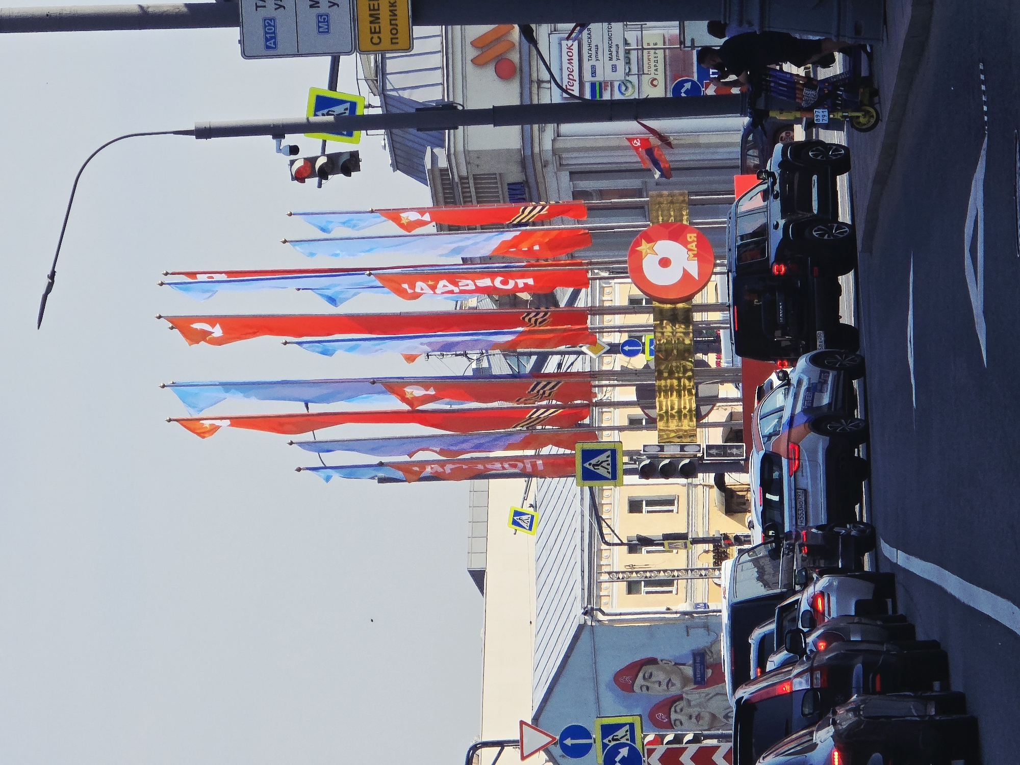 Here is a description of the image:
The image shows a busy street in Moscow, Russia. Several vehicles are visible on the right side of the image, including a dark SUV, a white car with blue and orange stripes, and several other cars and vans. Traffic lights and street signs are visible above the traffic. A person is riding a scooter on the sidewalk beside the traffic.
On the left side of the image, there are many red, white, and blue flags displayed above the street, and a large red sign with a white number "9" and the word "МАЯ" (May).