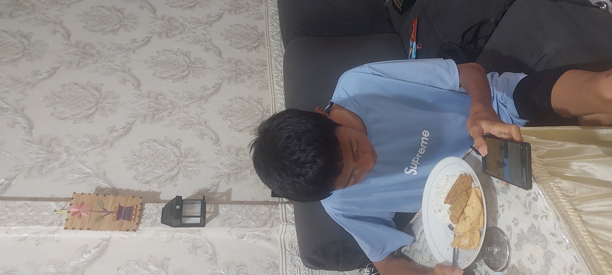 A young male, identified as Indonesian, sits at a table covered with a white lace tablecloth, in what appears to be a residential interior. He is wearing a light blue t-shirt with the word "Supreme" printed on it in white, and dark shorts. His gaze is directed downwards at a smartphone he holds in his right hand. In his left hand, he holds a fork, which is positioned over a white plate containing rice and what appears to be fried food items, likely tempeh and tofu. The setting includes a dark grey couch behind him and a wall adorned with ornate, textured wallpaper in a light neutral tone. On the wall, a small, dark rectangular object, possibly a solar-powered lamp, is mounted. To the left of the lamp, a rectangular craft made from wooden sticks depicts a pink flower in a brown vase. The overall lighting suggests an indoor environment, possibly during daylight hours. The provided image is unlikely to be from Jayapura, Indonesia as it is difficult to ascertain the specific location within a city without any identifying landmarks or context.