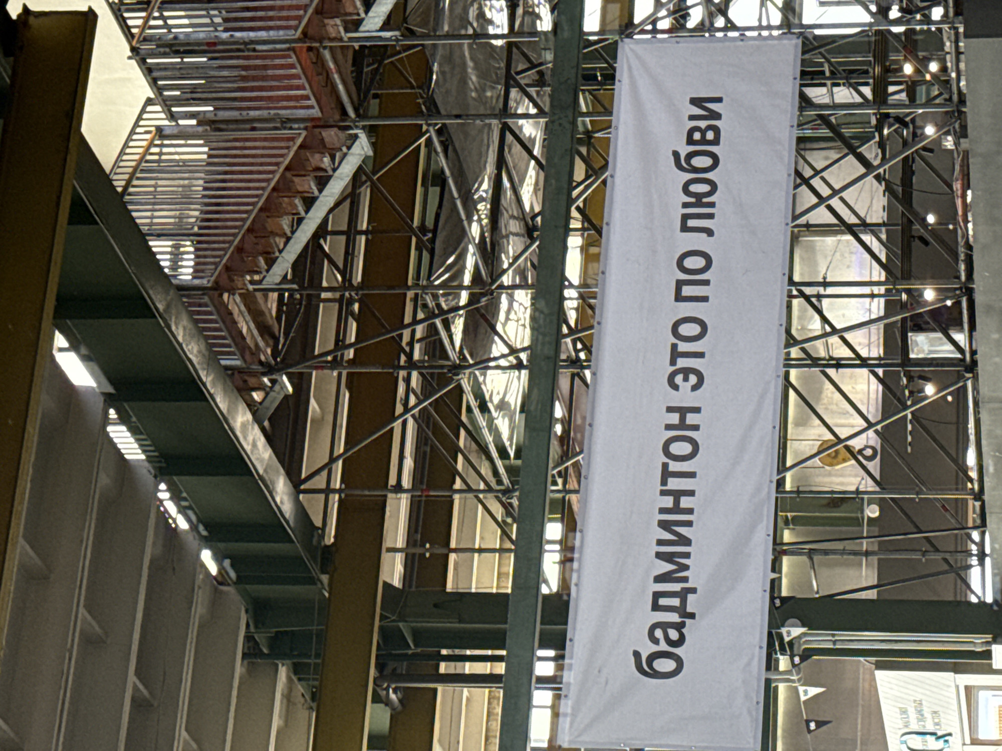 The image captures a white banner with black Cyrillic text, reading "бадминтон это по любви," which translates to "badminton is love." The banner is suspended in what appears to be an unfinished or under-construction indoor venue, possibly a sports complex or exhibition hall, given the exposed metal scaffolding and beams visible in the background. The lighting suggests an indoor environment with natural light filtering in from above. The banner itself is taut, indicating it is well-secured, with grommets visible along its top edge. The background is cluttered with construction materials and structural elements, including metal stairs and railings, suggesting a large, industrial-type space. The overall impression is of a work in progress, with the banner serving as a prominent, possibly promotional, display within the raw structure. The context of Saint Petersburg, Russia, is not evident from the visual elements alone.