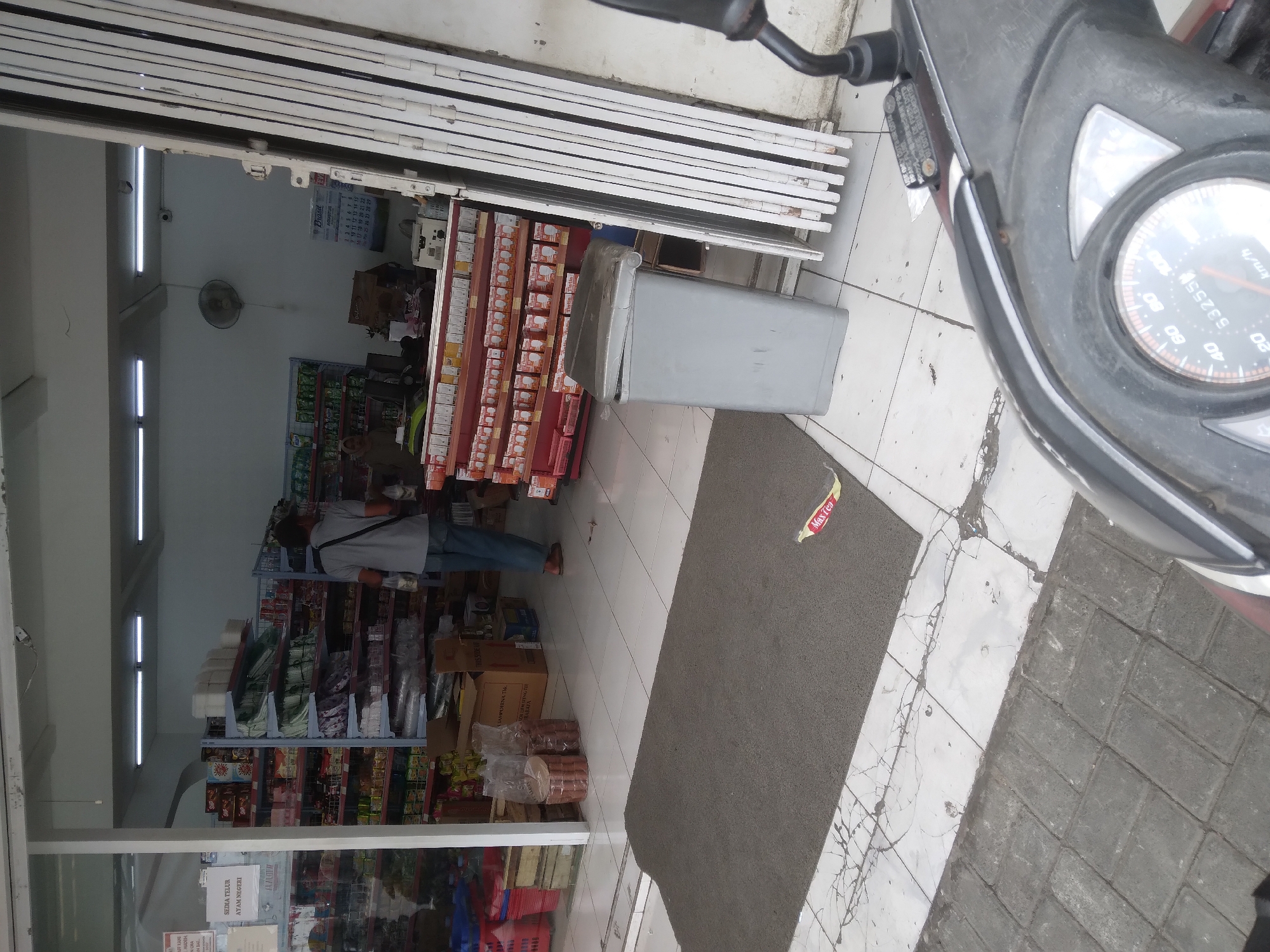 The image shows the entrance of a convenience store. Inside the store, shelves are stocked with various goods, including what appears to be food products and beverages. A person wearing a gray t-shirt and blue jeans is walking through the store, carrying a bag. In the foreground, there is a gray mat on the tiled floor, and a trash can is located near the entrance. A motorcycle is partially visible in the lower right corner of the image, suggesting the viewpoint is from someone on or near a motorcycle. The overall environment appears to be an indoor commercial space. Based on the context of the provided crops and the general appearance of the store, it is likely a typical convenience store found in Indonesia. The specific mention of "Berkoh, Indonesia" in the prompt suggests this location may be relevant, although there are no direct visual cues within the image to confirm this exact location.