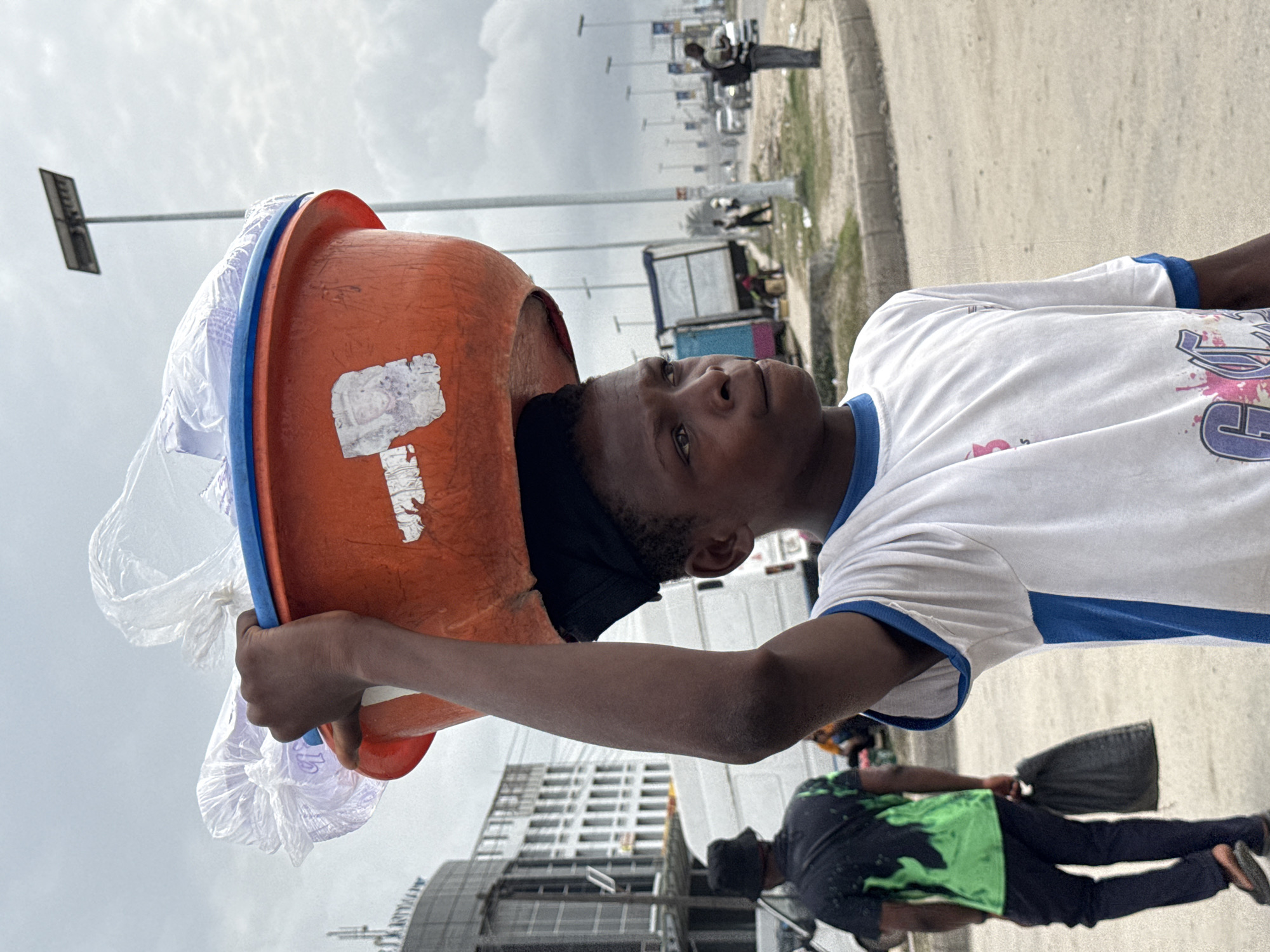 A person of African descent, with their head and shoulders inside a faded orange bucket with a blue rim, is visible in the foreground. The bucket is filled with a crinkled translucent material, potentially plastic packaging. The person's left arm is raised to hold the bucket, while the right arm is partially visible.
The person is wearing a white t-shirt with blue trim on the sleeves and collar, featuring a printed design. The individual is looking upward and away from the camera.
The background is an urban street scene. The setting appears to be in Lekki, Nigeria, evidenced by the architectural style of the building under construction in the lower frame.
A person wearing a dark shirt with green splotches is walking on the street carrying a dark bag. Further into the background are vehicles, lamp posts, and indistinct figures suggesting a busy street. The sky is overcast.