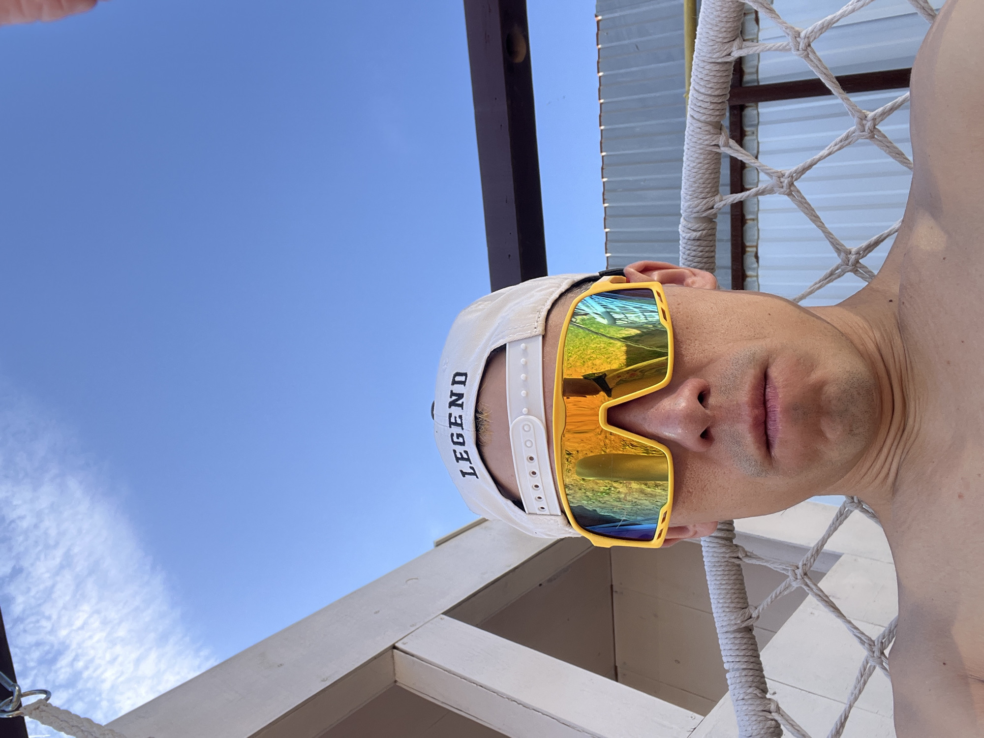 A man wears a white baseball cap with "LEGEND" printed on it and yellow sunglasses reflecting the sky. He is positioned beneath a structure with a white lattice frame, dark brown beams, and a metal roof. The sky is blue with sparse clouds. The man's shoulder is visible on the right side of the frame. The photo appears to be taken from a low angle, looking up. There is not enough information to determine the exact location in Chaikovsky, Russia.