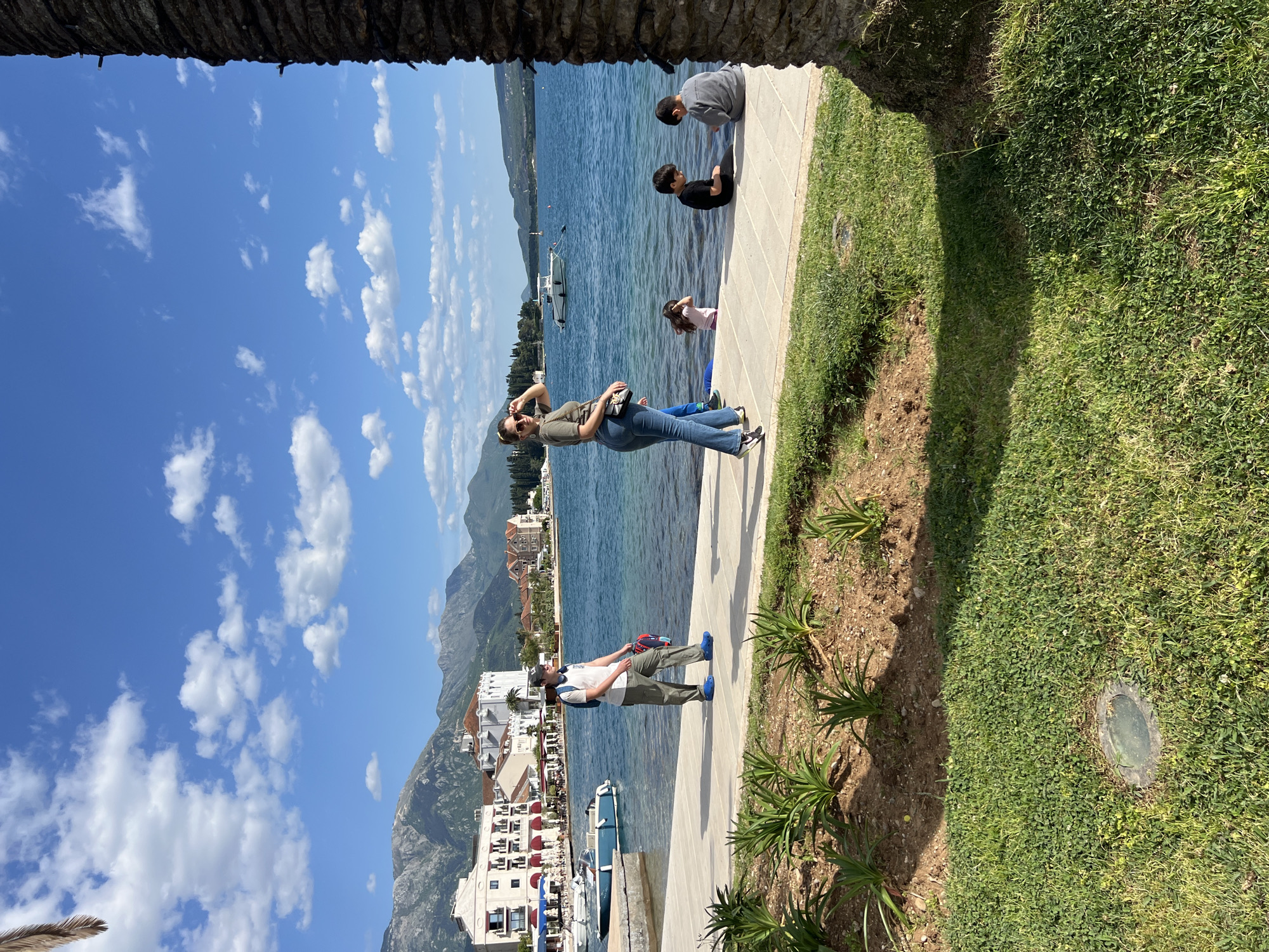The image, set in Tivat, Montenegro, captures a vibrant coastal scene on a sunny day. The clear blue sky, adorned with scattered white cumulus clouds, fills the upper portion of the frame. Below the sky, a serene body of water, likely a bay or sea, stretches across the middle and left side of the photograph. Distant mountains rise from the water's edge on the far side, and various white and light-colored buildings with red-tiled roofs dot the distant and closer coastlines, some with boats moored nearby.
In the mid-ground, a paved promenade runs parallel to the water's edge, separating it from a strip of green grass and small plants on the right. A large, textured palm tree trunk partially frames the foreground on the right and top edges.
Several people are enjoying the waterfront. In the mid-ground on the promenade, a woman wearing a dark olive top, blue jeans, sunglasses, and a black shoulder bag stands with a cell phone raised, seemingly capturing a photo or video of the scenery. Further down the promenade, towards the foreground, a man in a white t-shirt, olive pants, blue shoes, and a blue backpack stands, looking out towards the water and the distant town. Closer to the camera, two children are seated on the edge of the promenade, leaning over and looking into the calm water; one wears a gray top, the other a black t-shirt. A third child in a pink top is partially visible behind them, also near the water, and another person in blue pants is seen further back near the woman.
The bright sunlight casts distinct shadows on the grass and walkway, indicating a mid-morning or early afternoon setting. The scene conveys a peaceful and leisurely atmosphere. No visible text is present in the image.