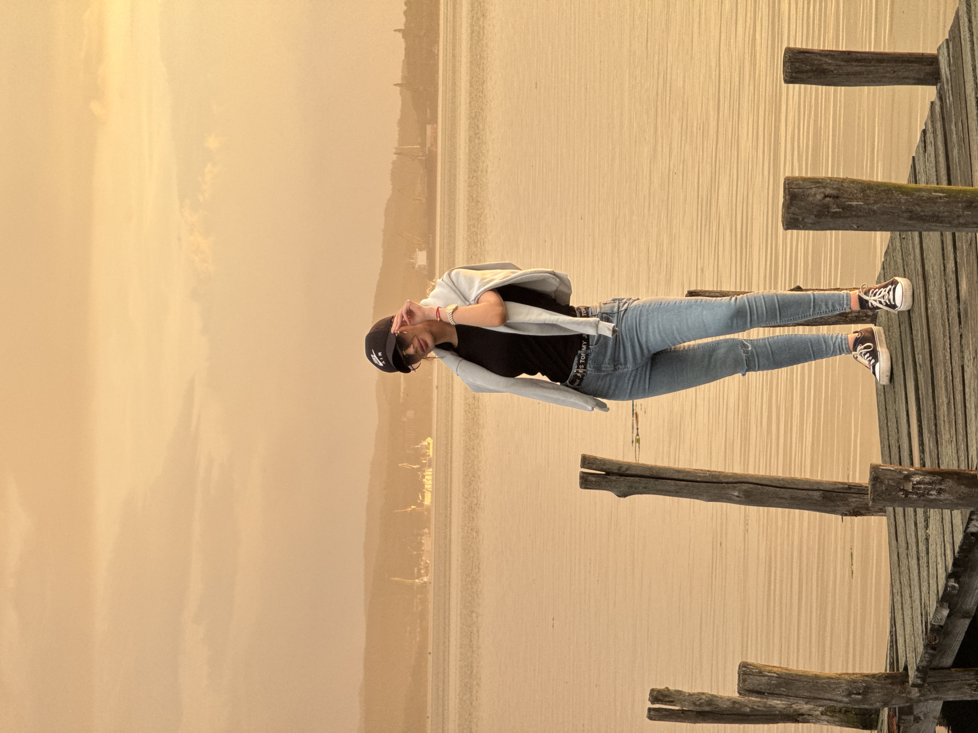 A person stands on a wooden dock in Казашко, Bulgaria. The person wears blue jeans, a black t-shirt, a light-colored jacket, a black baseball cap, and sneakers. The dock extends into a body of water. A hazy sky is visible in the background. The image is rotated 90 degrees clockwise.