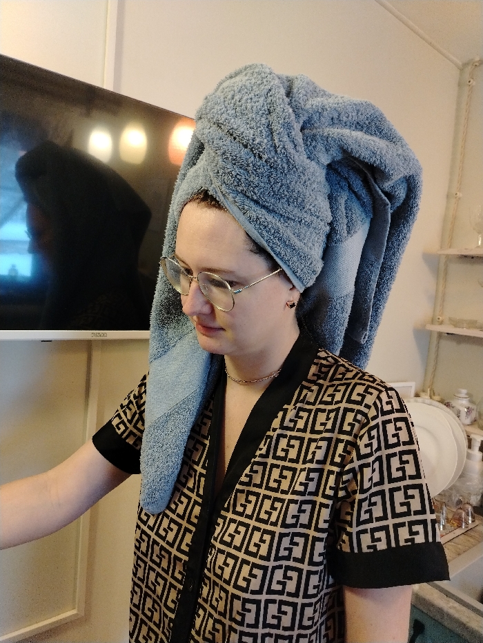 A woman wears glasses and a large, light blue towel turban on her head. She has a thin chain necklace and small earrings. She wears a patterned, tan-colored robe with black trim. A flat-screen TV hangs on the wall behind her, reflecting light. A white shelving unit holding dishes and toiletries is visible to her right.