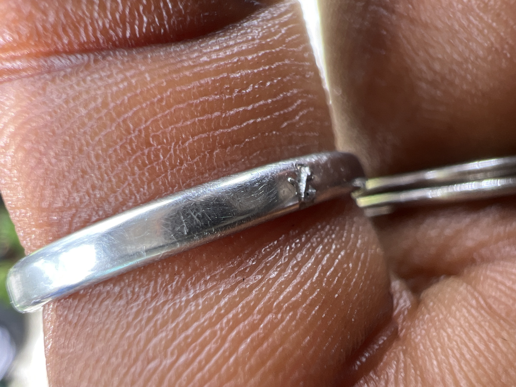 The media file is a close-up, macro photograph of a person's hand. The focus is on a silver ring that appears to be slightly damaged, with a small nick or crack visible on its surface. The skin of the hand is detailed, showing the natural lines and texture. The background is out of focus, suggesting a natural setting with some greenery visible, though not clearly identifiable. The lighting appears to be natural daylight, casting highlights on the ring and the skin. There is no visible text in the image. The image captures a moment of close examination of the ring on the hand. Given the location context of Yali, Nigeria, the skin tone suggests the person is likely of African descent. The overall impression is one of detail and texture, highlighting the intricate surface of both the human skin and the metallic object.