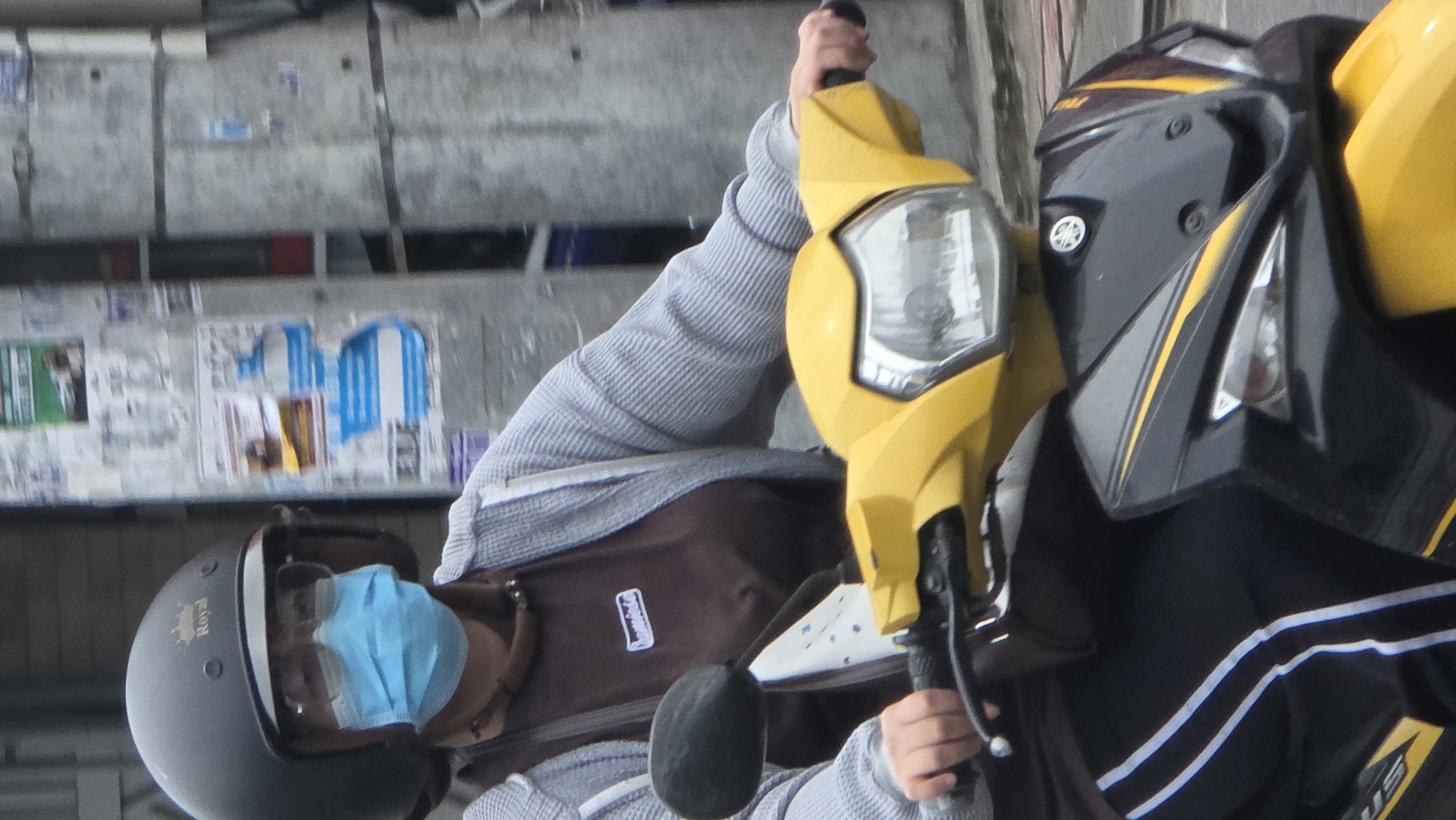 An eye-level shot captures a person seated on a yellow and black motorcycle in what appears to be an urban setting. The rider wears a dark grey open-face helmet with "Royal" written on its side in gold script. A light blue surgical face mask covers their lower face, and they are dressed in a light grey, long-sleeved, ribbed shirt with a dark brown bag or vest visible underneath. Their hands are gripping the handlebars of the motorcycle, which features a prominent yellow front section with a clear headlight. The side fairing of the motorcycle is black with yellow stripes and bears the "Yamaha" logo. Part of another similar motorcycle, also yellow and black, is visible to the right. In the background, a textured concrete wall is densely covered with numerous faded and torn posters and flyers, including patches of blue and white. The scene suggests the person is stationary, possibly waiting or stopped. The lighting indicates it is daytime, likely under overcast conditions or in a shaded area.