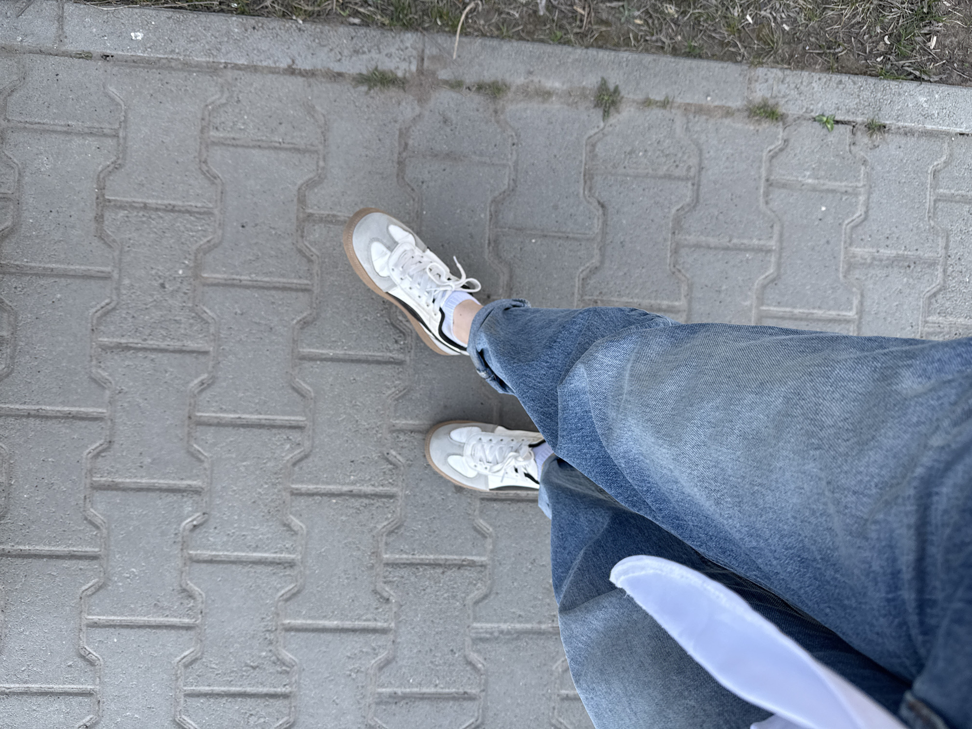A high-angle shot depicts a person's legs and feet standing on a paved surface constructed of gray, interlocking brick pavers. The person is wearing light wash denim jeans that are cuffed at the ankles, revealing white socks. Their footwear consists of white sneakers with tan-colored soles and black accent stripes. The pavers are arranged in a uniform pattern and show some minor wear and weathering. A patch of dirt with sparse vegetation borders the paved area on the right side of the frame. A white object, possibly paper, is visible near the bottom of the image partially obscured by the jeans. The location is an urban environment.