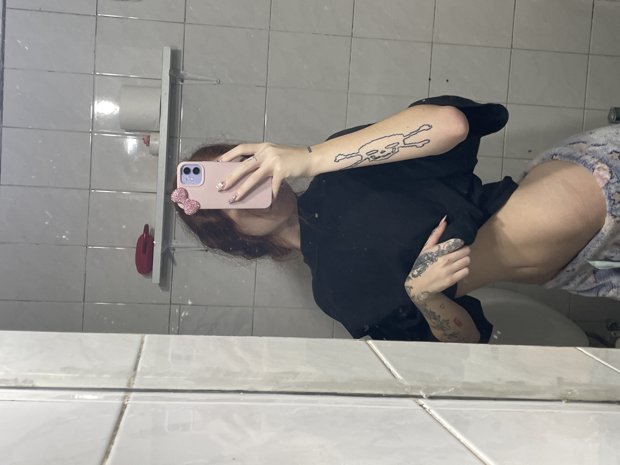 This image is a mirror selfie taken in a bathroom setting. A person is captured in the reflection, holding a light pink or lavender smartphone that largely obscures their face. The phone case features a decorative, glittery pink bow on its bottom left corner.
The person has dark brown or auburn hair visible around their ear and jawline. They are wearing a black short-sleeved top and patterned shorts, which appear to be light-colored with white or light-toned designs, possibly stars or abstract shapes. Both of the person's arms are tattooed. The left forearm displays a distinctive black outline tattoo in a pixelated style, depicting what looks like a dancing or jumping figure. The right arm and hand also bear several black ink tattoos with more intricate, possibly floral or ornamental patterns. Their fingernails are manicured, with some featuring glitter or decorative elements.
The bathroom features white or light grey tiled walls with darker grout lines. A roll of toilet paper is visible on a wall-mounted dispenser behind the person. A red object, possibly a cleaning tool, hangs on a white rack near the toilet paper. Part of a white porcelain fixture, likely a toilet bowl or the edge of a tub, is visible in the lower right portion of the mirror's reflection. The mirror itself appears to have some smudges or marks. The lighting is indoor, suggesting artificial light. No legible text is visible in the image. The location context provided is Buenos Aires, Argentina.