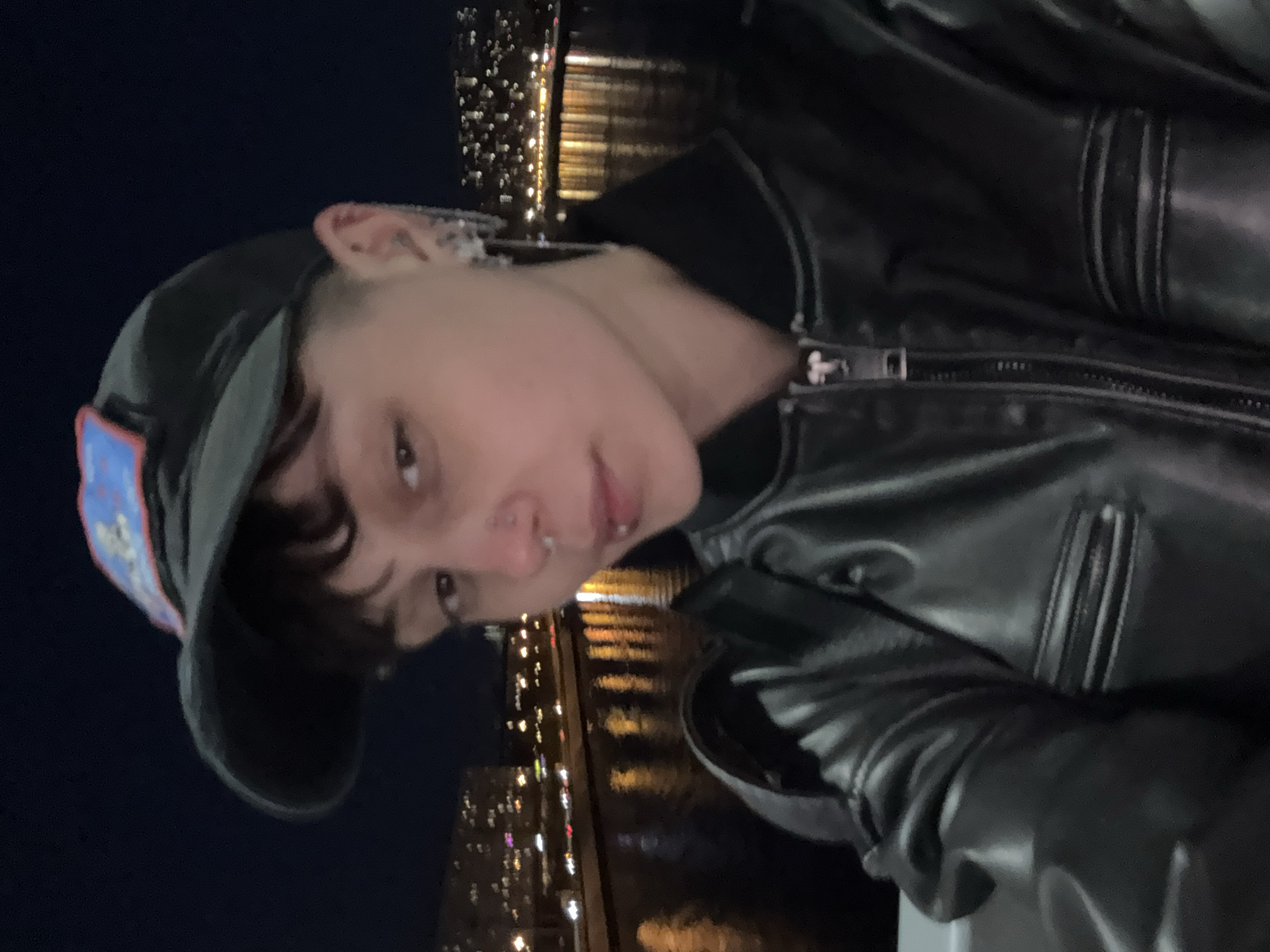A night-time portrait shows a young person in a dark outdoor setting, likely a city waterfront. The subject, positioned centrally, has short dark hair and looks directly at the camera with a neutral expression. They wear a dark baseball cap with a colorful patch on the side, featuring blue, red, and white elements (text or specific imagery on the patch is not discernible). A black leather jacket with a prominent zipper is worn, and the zipper pull appears to have a cross-like design. The person has multiple piercings, including a septum nose ring and possibly a lip piercing, as well as multiple earrings on their visible ear.
The background is dark with numerous blurred, sparkling lights reflecting on a body of water, suggesting a city skyline at night. The reflections create horizontal streaks of light on the water's surface. The setting implies a location next to a river or canal, possibly a bridge or embankment. Given the context of Saint Petersburg, Russia, this could be one of its iconic waterways. The overall mood is cool and urban, captured in a close-up, possibly a selfie. No legible text is visible in the image.