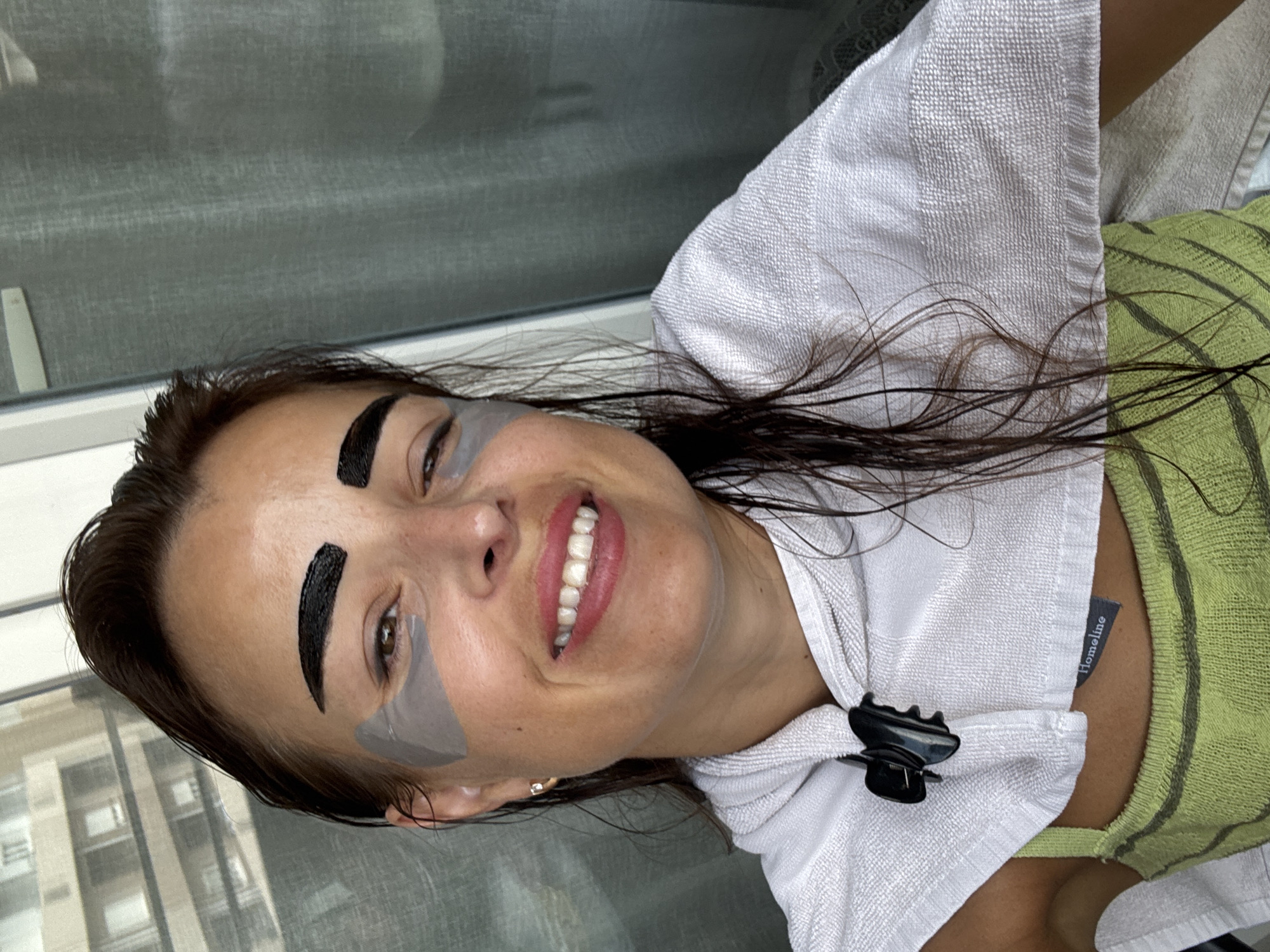 A woman with wet, dark brown hair, styled with a slight flip at the front, is shown from the chest up, facing the camera. She is smiling, revealing her teeth. She has dark, bold eyebrows and grey under-eye patches. A white towel is draped around her shoulders, secured with a black clip. Underneath the towel, she is wearing a light green tank top with dark horizontal stripes. The background consists of a blurred building with visible windows and a grey curtain. The overall impression is a selfie taken indoors, possibly in a bathroom or bedroom, given the grooming products and attire.