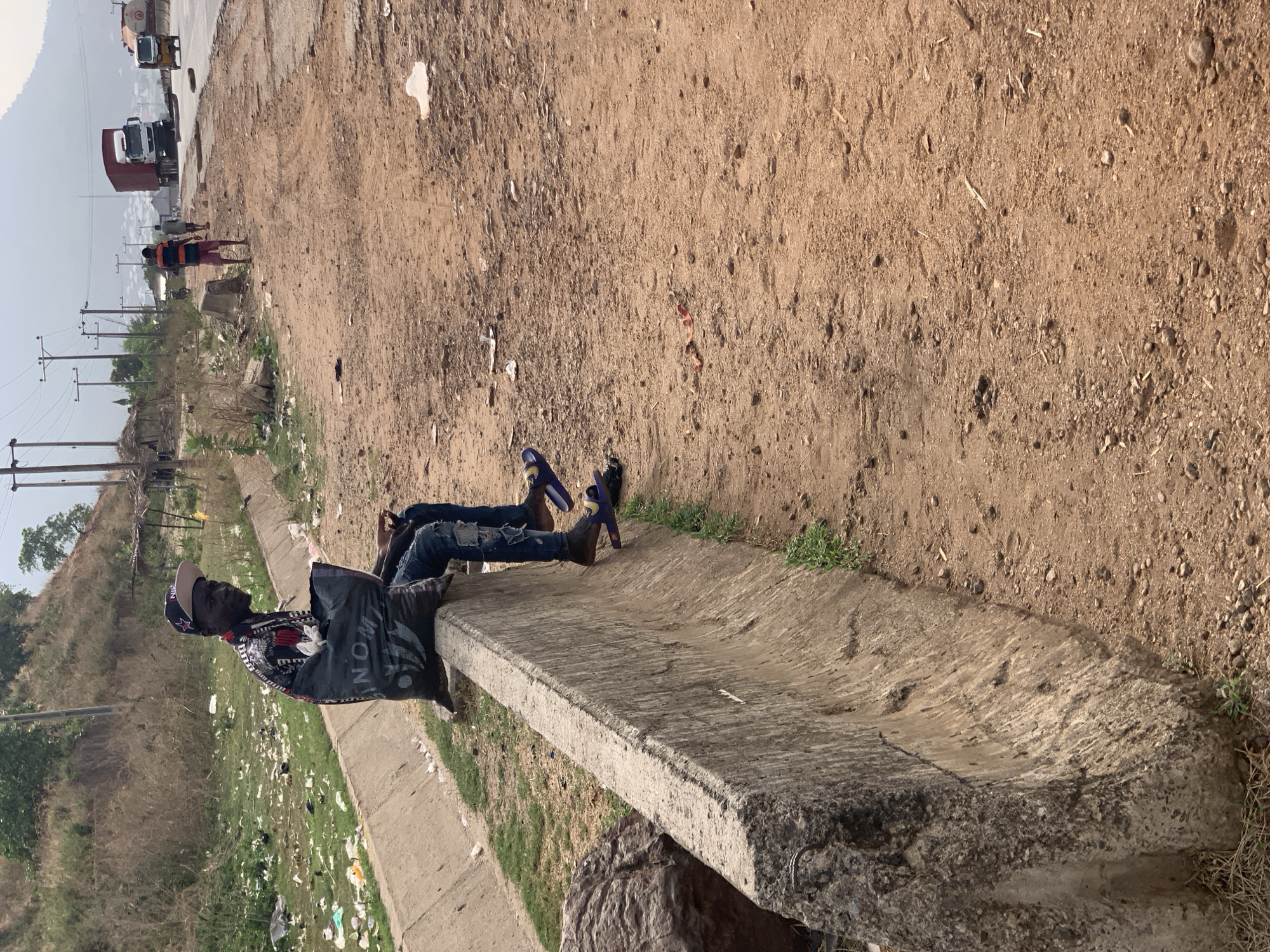 The image, taken in Lokoja, Nigeria, depicts an outdoor scene under bright daylight, likely late morning or afternoon, characterized by dry, hazy conditions. Two men are prominently featured, resting on a low, weathered concrete barrier that runs parallel to a dirt path.
In the foreground, one man is seated on the concrete barrier, facing towards the left side of the frame. He wears a patterned cap, a dark jacket with white and red patterns, and ripped blue jeans. The word "MON" is partially visible on his jacket. His left arm rests on his knee. Slightly behind him, another person's legs are visible, also resting on the barrier, wearing blue sandals and distressed jeans. The concrete barrier itself is wide and flat, appearing to be part of a drainage system, and shows signs of wear and tear. A large, dark rock or boulder is visible at the base of the barrier in the foreground.
The immediate surroundings feature a wide, unpaved dirt path to the right of the barrier, with scattered small stones and some debris. To the left of the barrier, a grassy embankment slopes downwards, heavily littered with various pieces of plastic, paper, and other waste. Beyond this, electrical poles with wires stretch into the distance, with sparse vegetation and some distant structures visible.
In the background, further along the dirt path, two more figures are seen walking away from the viewer. Behind them, a paved road is visible with large trucks, including one with a red container and another white cabin truck, suggesting a main thoroughfare or highway. The sky is bright and slightly overcast or hazy, contributing to the soft light. The overall setting is semi-urban or rural, marked by utility poles, unkempt areas, and ongoing vehicular traffic in the distance.