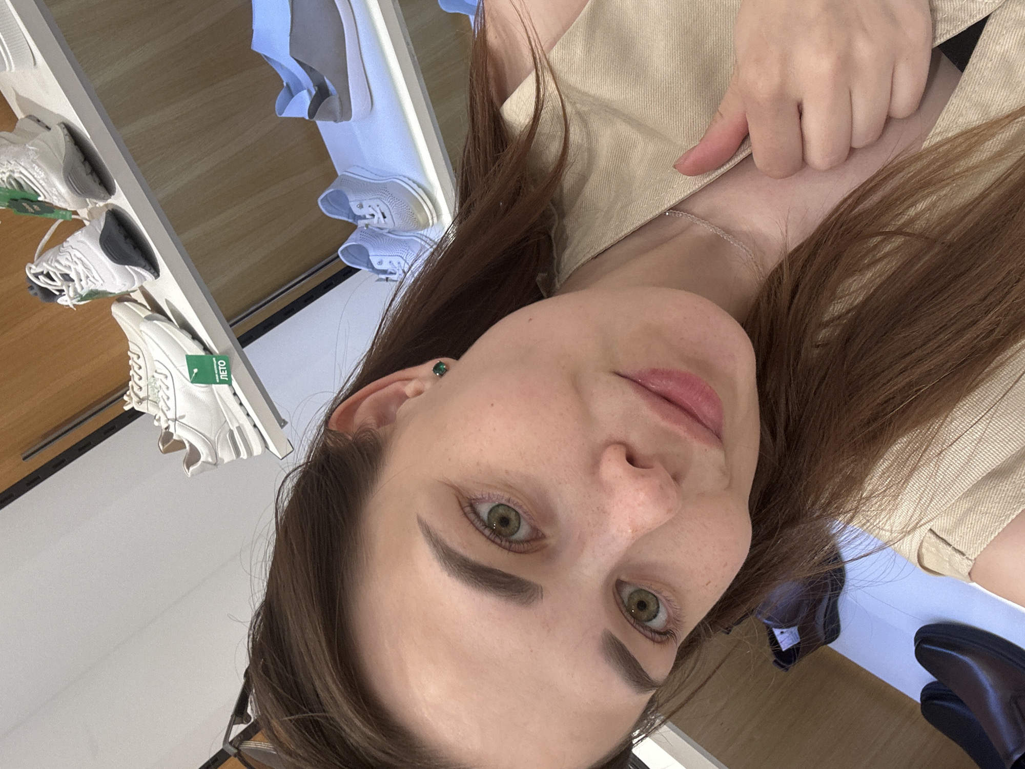 The image is a close-up selfie of a woman with fair skin, green eyes, and brown hair. She is wearing a beige shirt. A silver necklace is visible on her neck, and she has a green stud earring. The background shows a shoe store, with shelves displaying various white sneakers. A "ЛЕТО" (Summer) tag is visible on one of the shoes. The location is likely a store in Perm, Russia.
