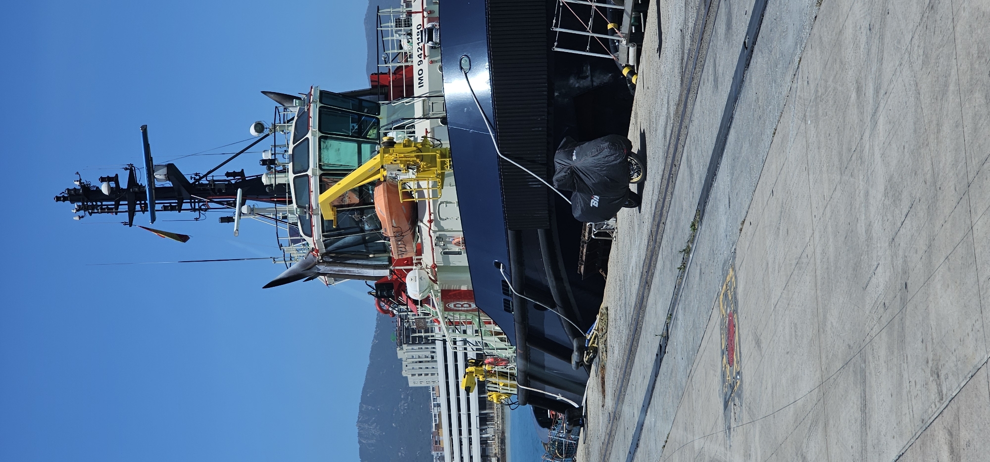 A large, dark-hulled vessel, likely a tugboat or offshore support vessel, is prominently docked alongside a concrete pier under a clear blue sky. The vessel features a multi-decked white and green superstructure with numerous windows. A yellow-colored crane with an orange life raft attached extends from the side of the superstructure. A complex mast with an array of antennas, navigation equipment, and lights rises high above the deck. The vessel's International Maritime Organization (IMO) number, "IMO 942-1190", is visible in white lettering on its hull, along with a partial letter "B" lower down.
On the concrete dock, a motorcycle covered in a black protective tarp is parked. The word "Bik_" is visible on the cover. Mooring lines from the vessel secure it to bollards on the pier. The dock surface shows concrete slabs, expansion joints, and faint tire marks. Metal rails are embedded into the concrete, running parallel to the ship, and a painted red and yellow rectangular marking is visible further along the dock.
In the background, beyond the vessel and the water, a cityscape with white and gray multi-story buildings stretches towards the base of imposing, rugged mountains. The scene depicts a bright, sunny day, with strong sunlight casting distinct shadows. The overall setting is a working port environment in Algeciras, Spain.