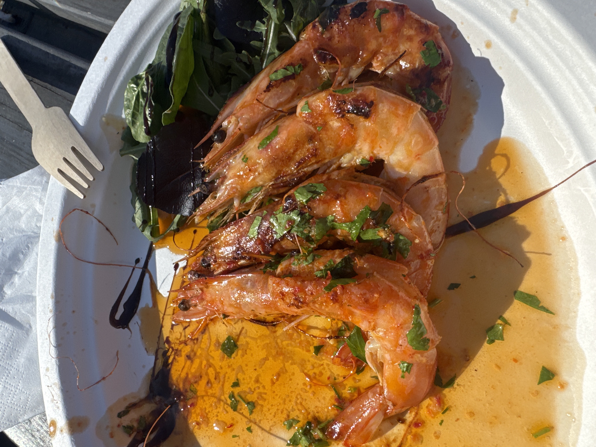 The image depicts a plate of grilled shrimp served with greens and a wooden fork. The shrimp are arranged in a stack, coated with what appears to be a glaze or sauce and sprinkled with herbs. The greens include arugula and what might be beet greens. The plate is white and appears to be made of paper. The setting seems to be outdoors, possibly on a picnic table, as suggested by the wooden surface visible at the left. There is no clear evidence to confirm that it is specifically in Epping Forest, United Kingdom.