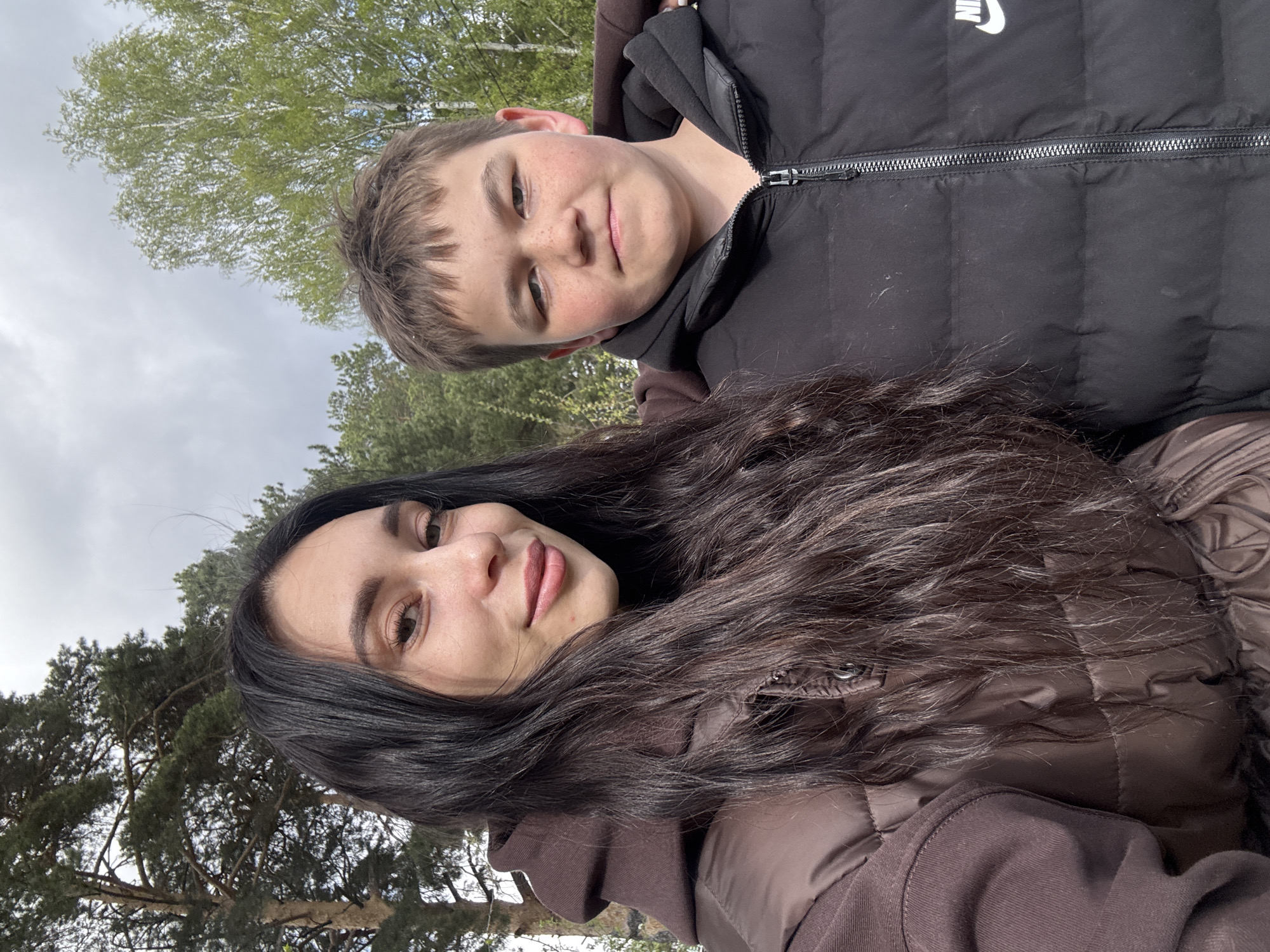 Two individuals are visible: a young male and a female. The male, fair-skinned with freckles, is wearing a black padded jacket with a white Nike logo. The female has dark hair and is wearing a brown padded jacket. They appear to be standing outdoors in a wooded area, with trees and an overcast sky visible in the background. The location is likely in a natural setting, possibly a park. The location in the city of Chaikovsky, Russia, is not discernible.