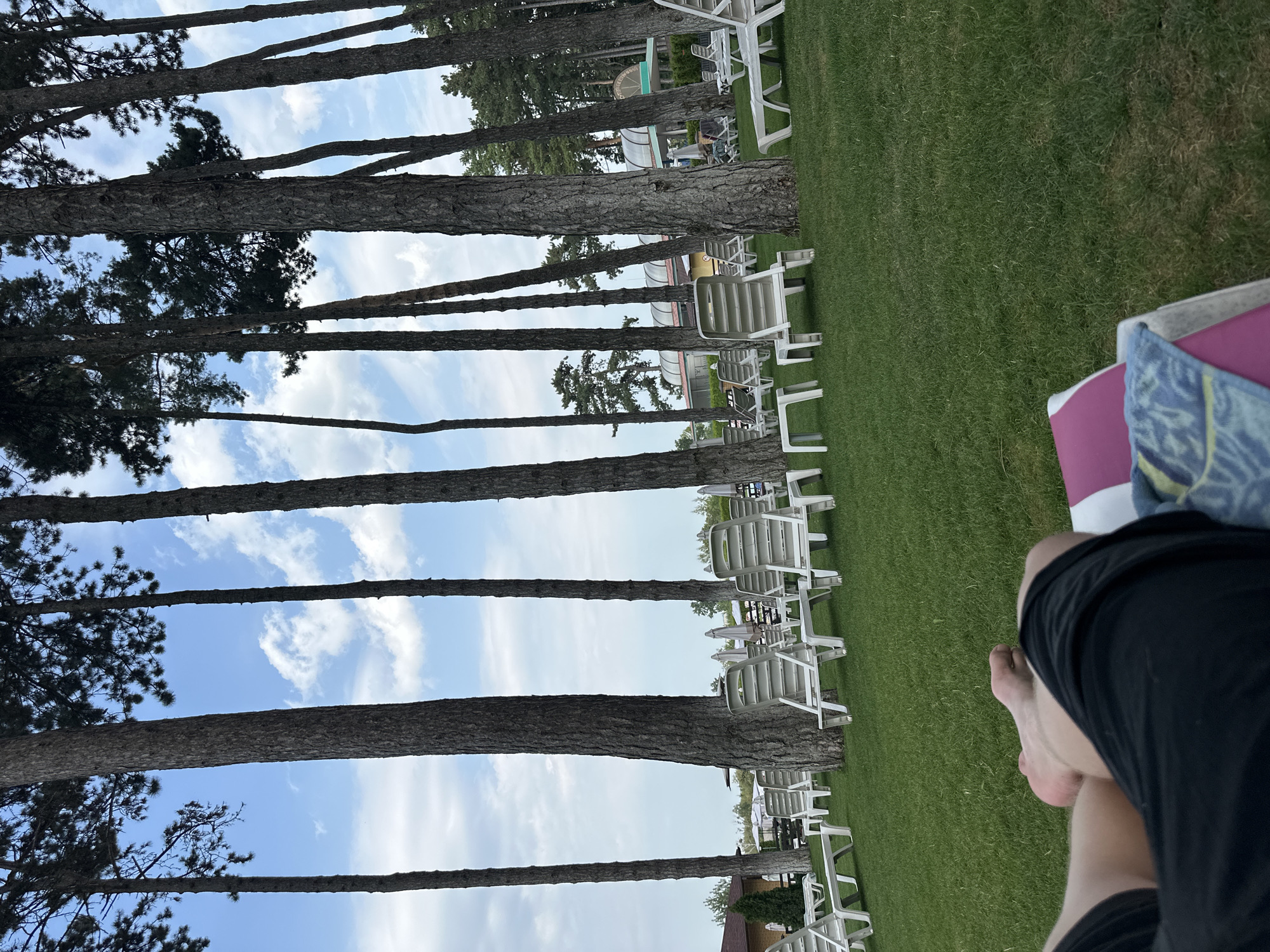 The image captures a sun-drenched scene at a resort in Khmilnyk, Ukraine. Tall, slender pine trees stand sentinel over a meticulously manicured lawn, where rows of empty white lounge chairs await relaxation. In the foreground, a person reclines on a lounge chair, their legs crossed, dressed in black shorts. The sky above is a canvas of blue, adorned with scattered white clouds. The resort's facilities, including a building with a curved roof and what appears to be a swimming pool area, are visible in the background. The overall impression is one of serene tranquility, a perfect day for leisure.