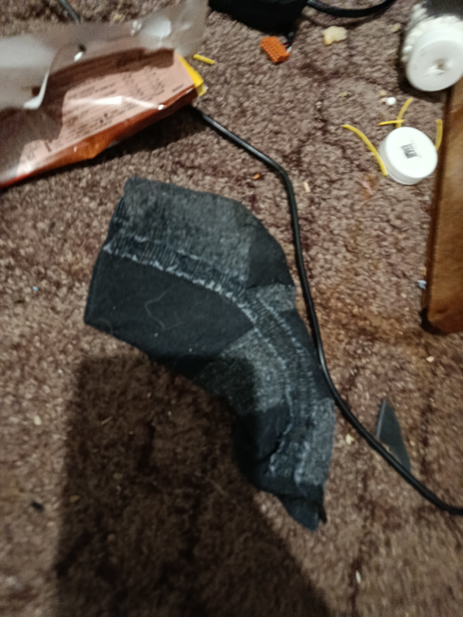 A dark grey sock lies on a brown, fuzzy carpet. The sock is folded and appears to be slightly damp or heavily used. A black cord snakes across the carpet, passing near the sock and disappearing out of frame. In the background, partially visible, is a torn open snack wrapper with orange and pink packaging. To the right, there are two small white spools with yellow wires attached. The lighting suggests an indoor setting, possibly in the evening or under artificial light. The overall scene is cluttered and appears to be a casual, unorganized living space. No people are visible in the frame. No text is legible.