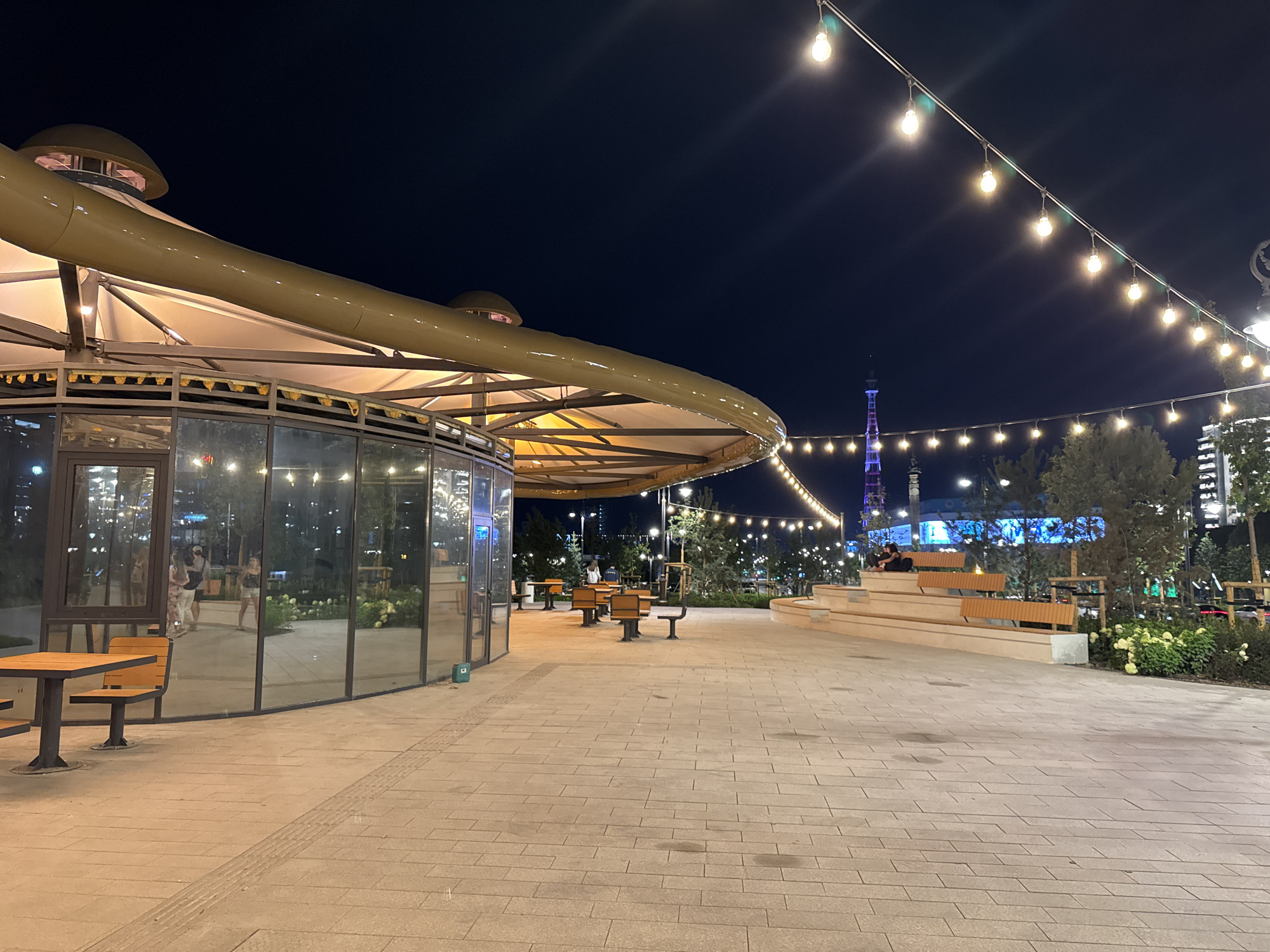 The image depicts an outdoor plaza at night in Almaty, Kazakhstan. In the foreground, there is a paved walkway with large rectangular tiles. To the left, a modern building with glass walls and a distinctive yellow curved roof structure is visible. Inside the building, reflections show several people. In front of the building, there are outdoor tables and chairs.
Further into the plaza, to the right, a tiered seating area with wooden benches is present. Beyond the seating area, there are trees and shrubs, and in the distance, a tall tower with lights is visible. Strings of decorative lights are strung across the plaza, illuminating the scene. The overall environment suggests a public gathering space or entertainment area, possibly a park or a newly developed urban zone.