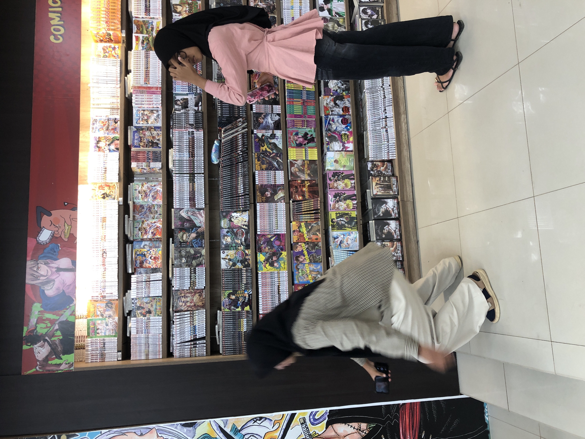 Two individuals wearing hijabs are browsing manga on shelves inside a comic book store, identified as "COMIC" from a sign on the wall. The store is located in Banda Aceh, Indonesia.
The person on the left, partially obscured by motion blur, is crouching and holding a mobile phone. They are wearing a light-colored, striped long-sleeved shirt and loose, light-colored pants, with sandals.
The person on the right is standing and looking towards the shelves. They are wearing a pink peplum-style top and dark pants, with black sandals. Their right hand is raised to their face.
The shelves are stocked with numerous volumes of manga, with colorful covers visible. A large mural featuring anime characters is displayed above the manga shelves. The floor is tiled with large, white square tiles. The overall lighting suggests an indoor retail environment.