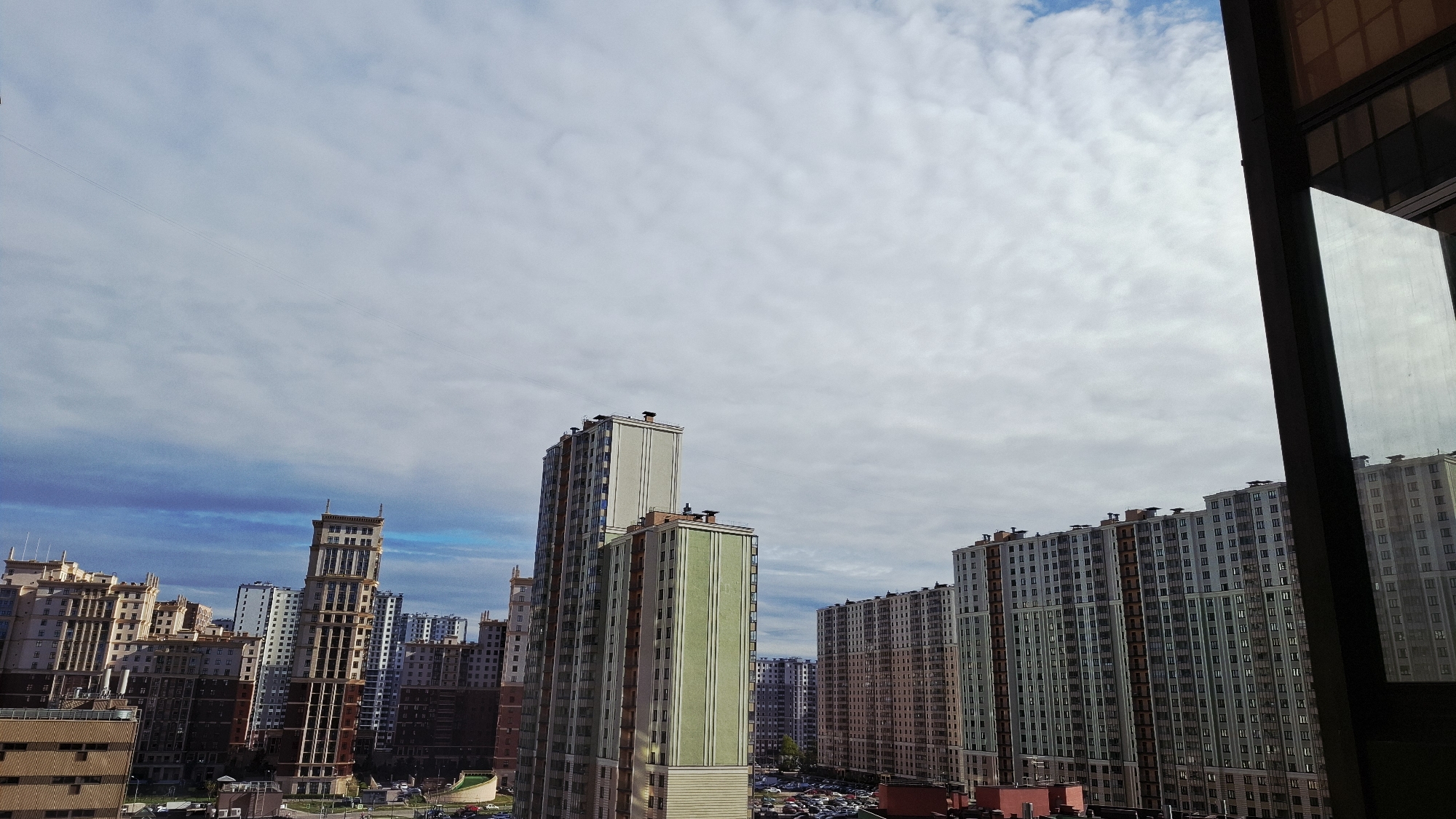 This image shows an urban view in Saint Petersburg, Russia, featuring multiple high-rise residential buildings under a partly cloudy sky. A window frame is visible in the right foreground, suggesting the photograph was taken from inside a building. The buildings vary in design, with some featuring modern facades and others exhibiting more classical architectural elements. There's a large parking area visible between the buildings, filled with cars. The lighting suggests it is daytime. No people are clearly visible in the scene. The overall environment appears to be a dense urban area.