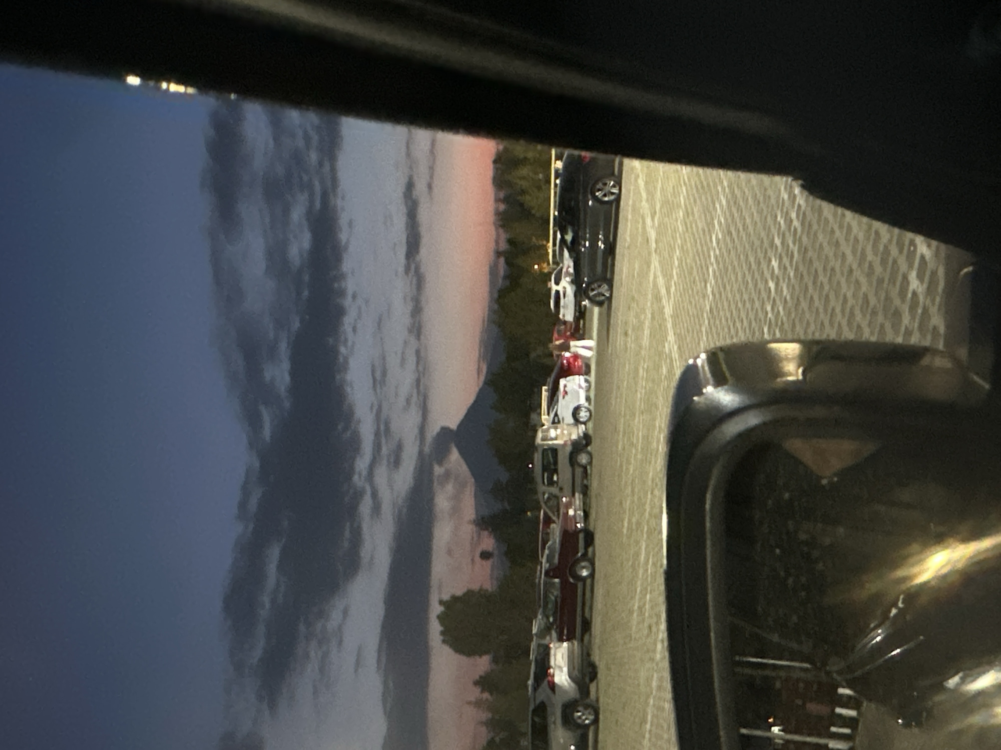 The image, taken from within a vehicle, captures a parking lot scene at either dusk or dawn in San Andrés Cholula, Mexico. The sky transitions from a deep blue at the top to soft orange and pink hues near the horizon, indicative of sunrise or sunset. Large, dark clouds are prominent against the colorful sky.
In the distance, the distinct silhouette of a large mountain or volcano, likely Popocatépetl, stands against the horizon. Below it, a line of parked cars of various models and colors, including black, white, and maroon, fills a paved parking lot. The pavement features a patterned texture. Trees and bushes are visible along the mid-ground, partially obscuring the base of the distant landform.
The foreground is dominated by a dark frame, presumably the vehicle's window frame, and a significant portion of a side-view mirror, which reflects parts of the parking lot and some distant lights. Small, bright lights can be seen on the distant horizon, possibly from a town or structures on a ridge. There are no visible people or discernible text.