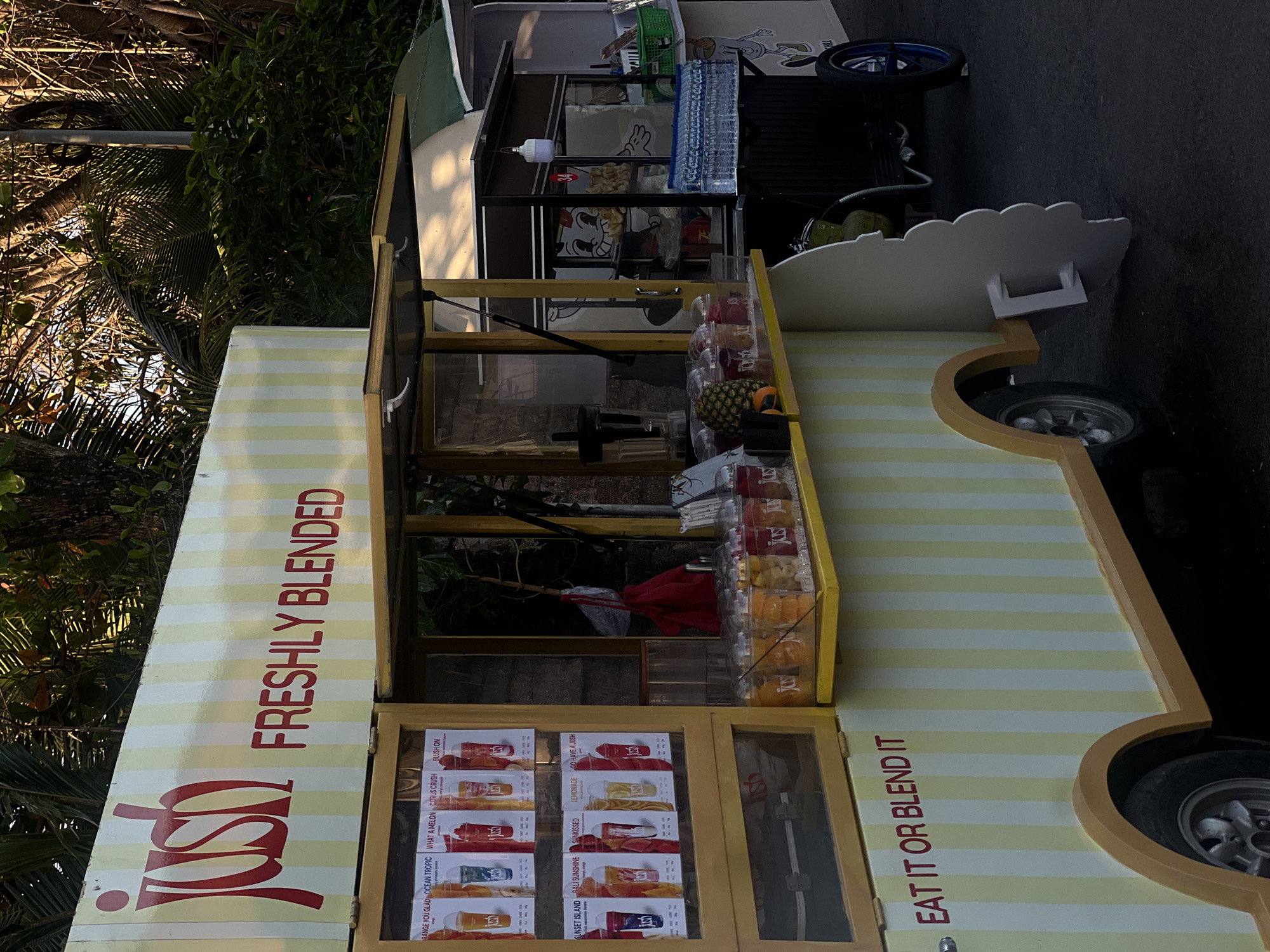 The image shows a juice stand labeled "JUSH FRESHLY BLENDED" in North Kuta, Indonesia. The stand is painted with yellow and white stripes and has a menu board displaying various juice options. Several clear plastic cups filled with colorful liquids and fruit are visible on the counter. In the background, there are trees and other food carts. The lighting suggests it is daytime, possibly late afternoon, given the warm light illuminating the scene. The overall scene depicts a vibrant and casual street food environment.