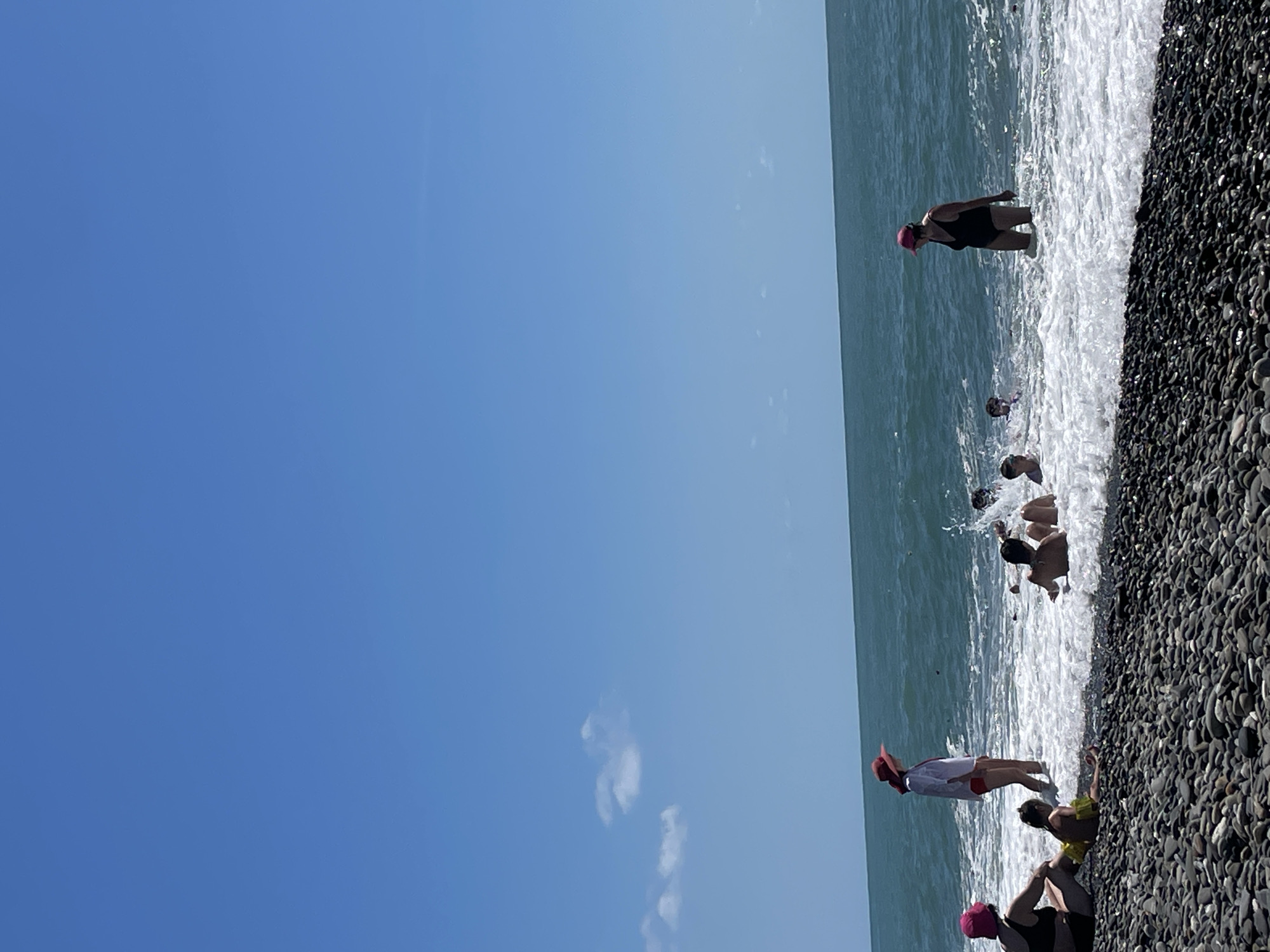 This is a photograph of a beach scene in Sochi, Russia, on a clear, sunny day. The foreground is composed of dark, smooth pebbles. The ocean, a deep blue-green, occupies the middle ground, with white waves breaking on the shore. In the water, several people are visible. On the left side of the frame, two women and a child are sitting on the pebbles, with the child wearing yellow flotation devices. Another woman in a white shirt and red hat is standing in the shallow water. Further out in the water, several individuals, including children, are playing and splashing. On the right side of the frame, a woman in a black swimsuit and pink hat is standing in the water. The sky is a bright, cloudless blue, with a few wispy white clouds visible near the horizon.