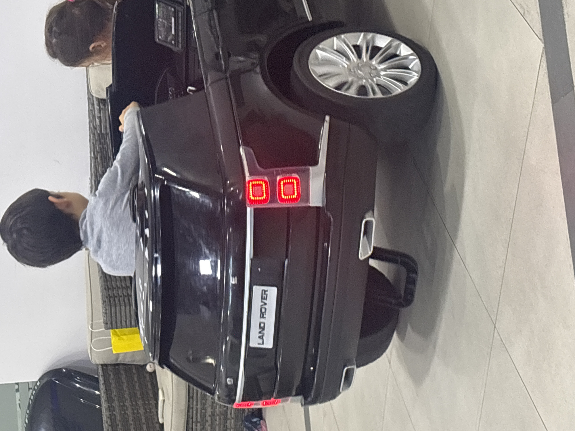 A black, toy electric Range Rover is parked indoors on a tiled floor, with the rear of the vehicle prominently displayed. Two young children are seated inside the toy car. A boy with dark hair is in the driver's seat, facing forward, and a girl with dark hair pulled back with a pink tie is partially visible in the passenger seat. The toy car features realistic details, including working red taillights, chrome accents, and a license plate that reads "LAND ROVER". The environment appears to be an indoor play area or a showroom, with a glimpse of outdoor furniture with cushions and a yellow shopping bag visible in the background. The lighting suggests an indoor setting. The scene is devoid of any overt signs of distress or unusual activity.