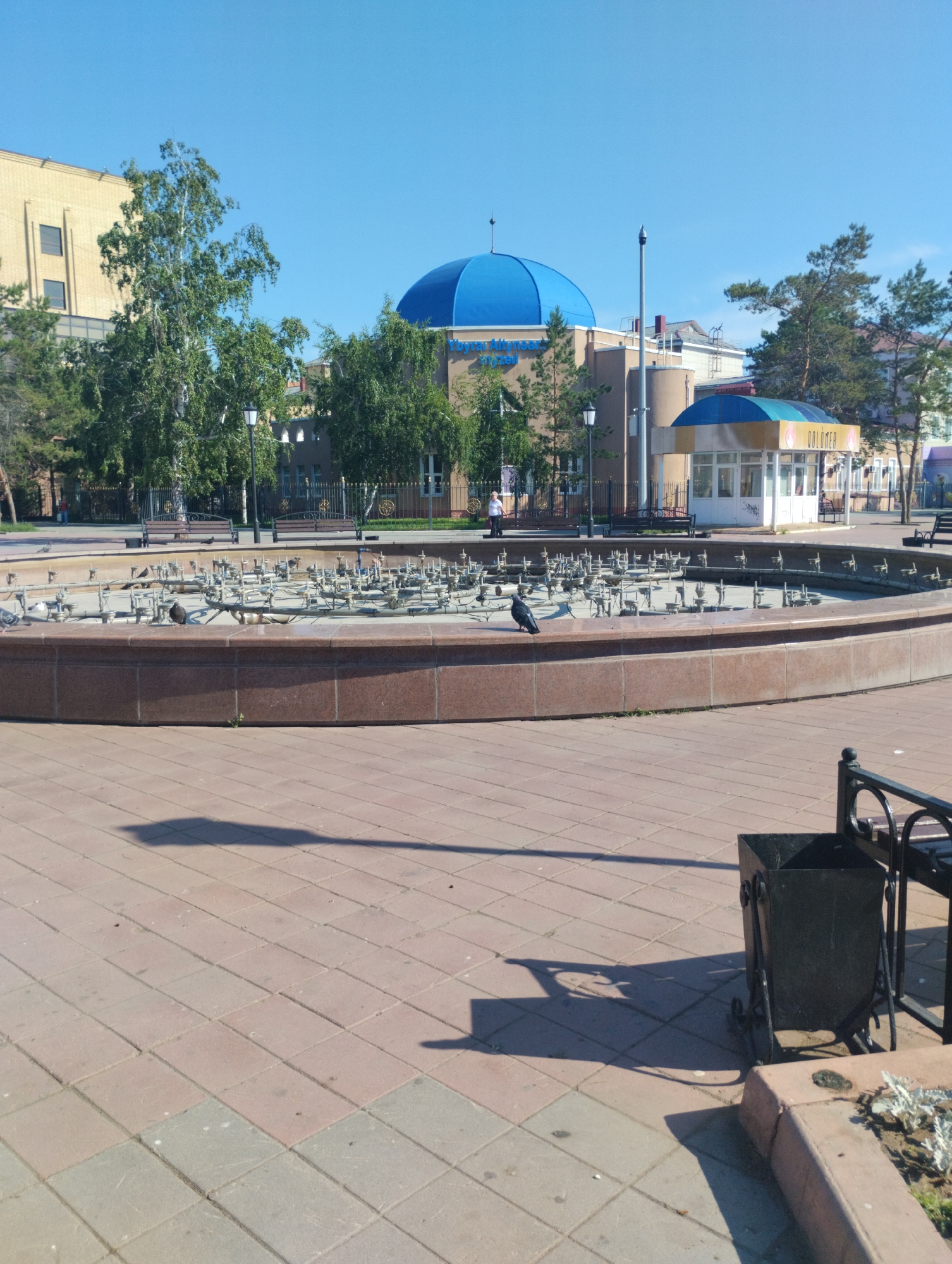 This image captures a public square or park in Kostanay, Kazakhstan, located directly in front of the Kostanay Regional Universal Scientific Library named after L.N. Tolstoy. The library is identifiable by the prominent bright blue dome and the visible text "Орталық кітапхана" (Central Library) on its facade, situated in the central background.
In the foreground, a large, circular, dry fountain with a maroon-colored granite rim is central. Its basin is empty, revealing numerous metal nozzles and spray mechanisms arranged in concentric circles. A single dark gray pigeon is perched on the fountain's rim, facing left, with several other pigeons visible within the empty basin and on the surrounding paved area.
The ground is uniformly paved with square tiles in varying shades of reddish-brown and gray. Long, distinct shadows, including that of a lamppost, stretch across the pavement from the upper right to the lower left, indicating bright sunlight. To the immediate lower right, a black wrought-iron trash receptacle is partially visible.
In the midground, beyond the fountain, several dark-colored public benches and black lampposts are positioned along paved walkways. The area is bordered by green trees, including tall, slender deciduous trees and coniferous evergreens. A metal fence with decorative elements separates the paved area from a grassy section further back.
In the background, to the left, a large, light-yellow, multi-story building with multiple dark windows stands. To the right of the library, a small, white, single-story kiosk with a yellow and blue arched awning is visible. The word "DOLOMER" (or DOLOМЕР) is displayed on its front signage. A lone person in light attire is visible standing in the distance near a fence to the left of the kiosk. The sky overhead is clear and bright blue.