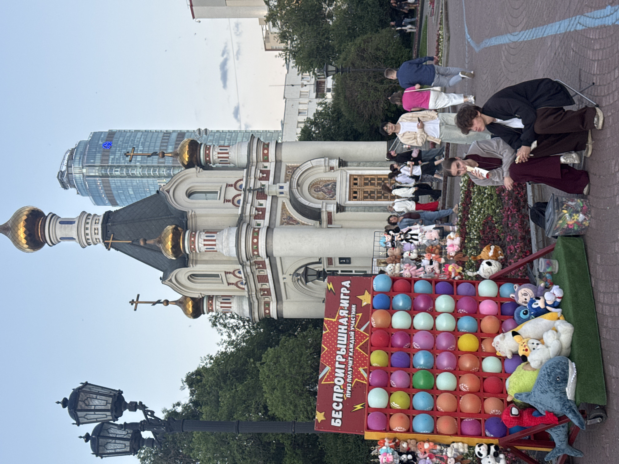 The image captures an outdoor scene in Yekaterinburg, Russia, likely in a public square or park. In the foreground, a carnival game stand is set up, featuring a large board filled with colorful balloons. Above the balloons, a red banner displays Russian text that translates to "NO-LOSS GAME, every participant gets a prize." Various stuffed animals are displayed around the game, serving as prizes.
To the left of the game stand, a vintage-style lamppost with two lamps is visible. In the background, a small, ornate church with golden domes stands prominently. Behind the church, a modern, tall glass skyscraper dominates the skyline, displaying a blue logo.
Several people are present in the scene. Two young individuals are seated on chairs in the mid-ground, appearing to be eating. Other people are walking in the background, suggesting a public gathering or event. The overall environment suggests a pleasant day, possibly in the late afternoon or early evening, with a mix of historical and contemporary architecture.