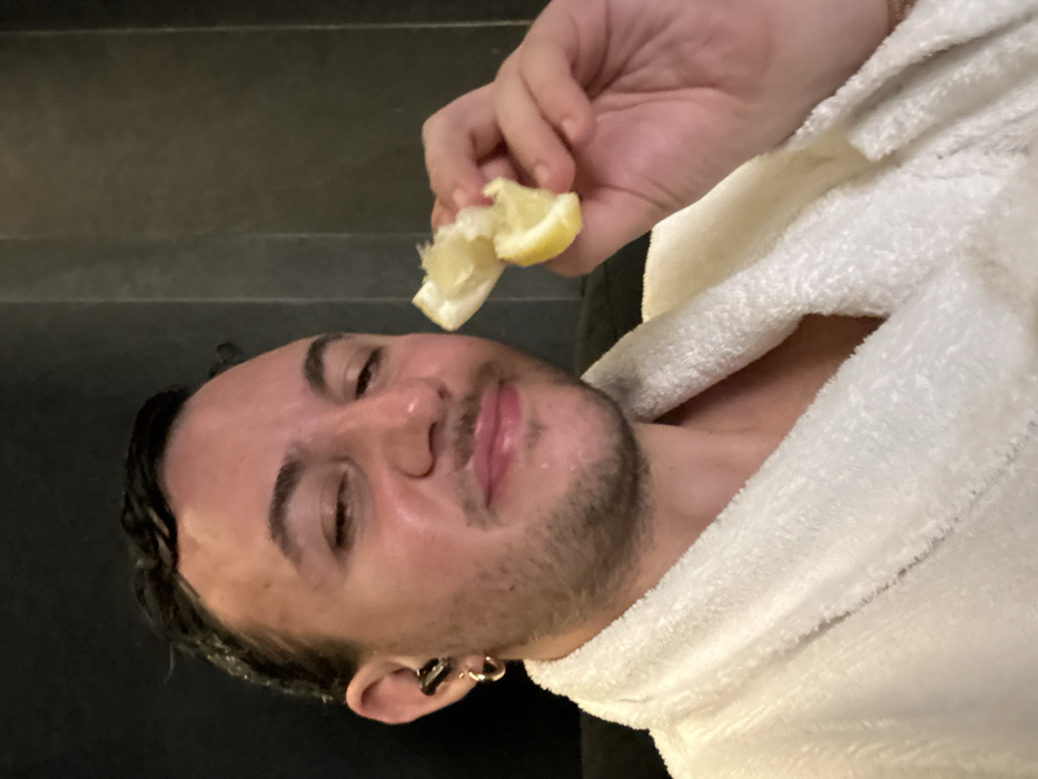 A male individual, appearing to be in his 20s or 30s, is depicted from the chest up in an indoor, dimly lit environment. His dark hair is wet and slicked back, and he has a mustache with light facial stubble. His skin appears moist. He is wearing a white terry cloth bathrobe. A small gold hoop earring is visible in his left earlobe, and a black earbud is in his left ear canal. He holds a partially squeezed or torn yellow lemon in his right hand, positioned near his mouth. His eyes are slightly squinted, and his lips are pursed in a slight smile. The background is composed of dark, vertically paneled walls. The image provides no discernable visual information to determine a specific location within Paris, France.