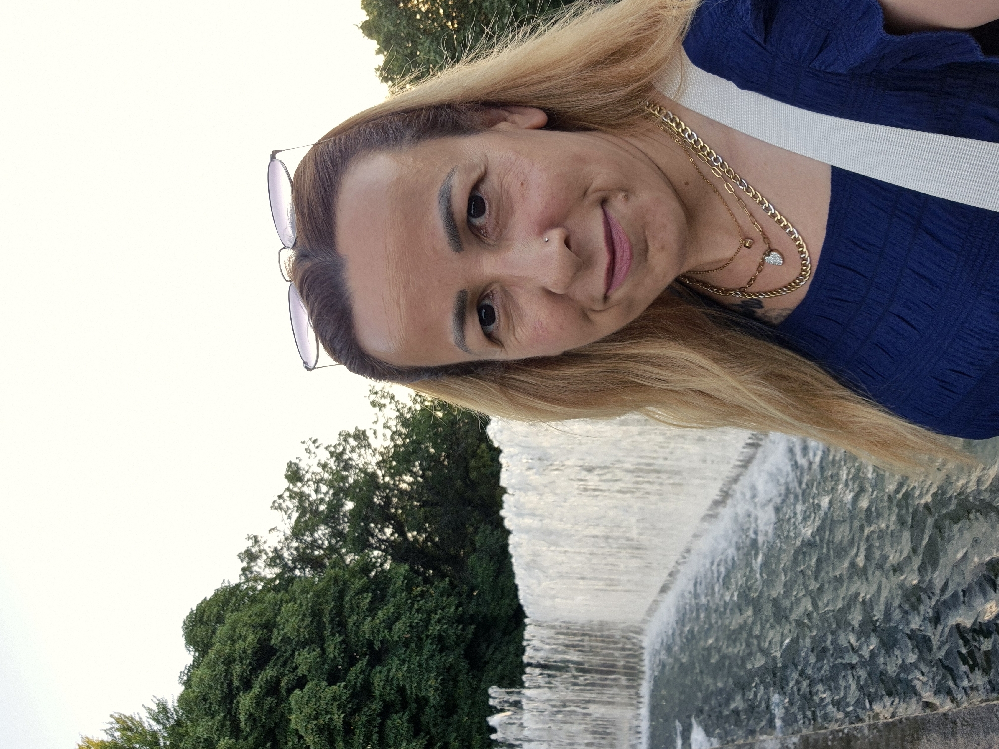 A woman with blonde hair and a blue top is posing for a selfie in front of a fountain. She is wearing multiple necklaces and sunglasses on her head. The fountain is long and wide, with water cascading down its side. Trees can be seen in the background. The lighting suggests it is daytime, possibly late afternoon or early evening.