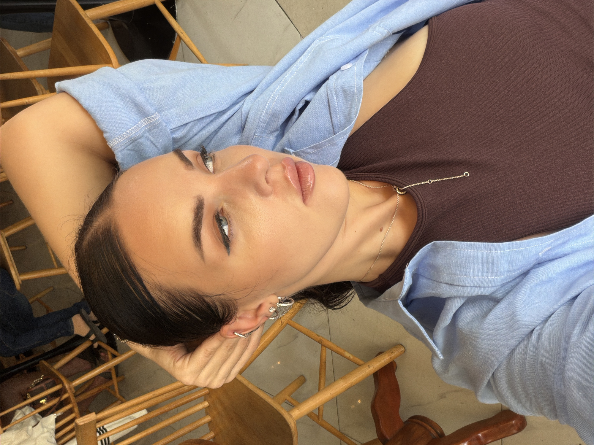 A young woman is lying in a chair. She has dark hair pulled back and is wearing a brown ribbed top and a light blue button-down shirt. She is looking up and to the left. She has multiple earrings in her left ear. A silver necklace is visible around her neck. The background consists of wooden chairs and a tiled floor. The location is Nha Trang Ward, Vietnam.