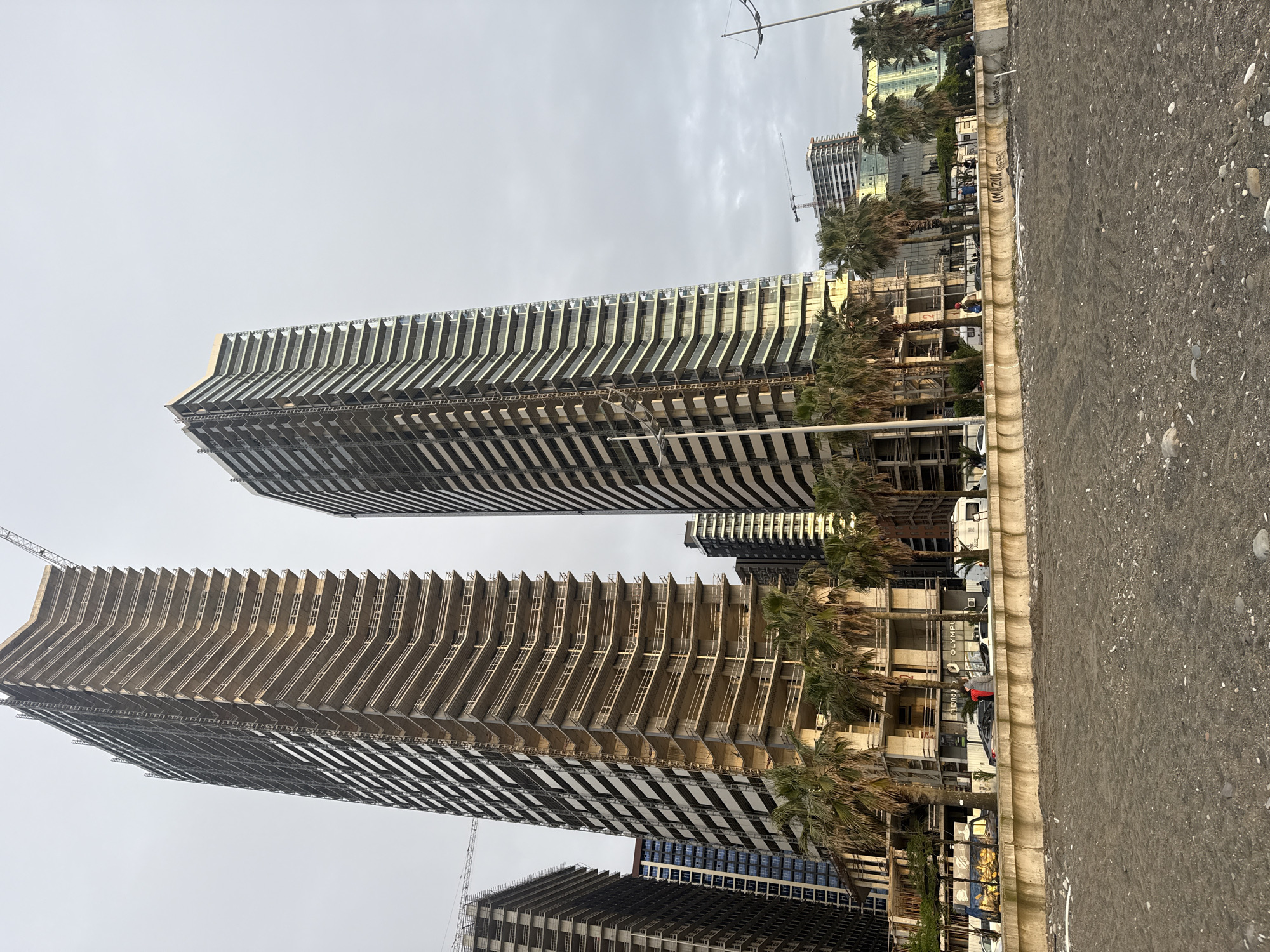 The image shows two tall, modern buildings under construction in Batumi, Georgia. The buildings have a distinctive design with repeating horizontal balconies. Palm trees line the street between the buildings and a sidewalk, leading to a beach. Construction cranes are visible, indicating ongoing development. The sky is overcast. A street with parked vehicles runs alongside the buildings. A shop called “Olympus” is visible at the street level.