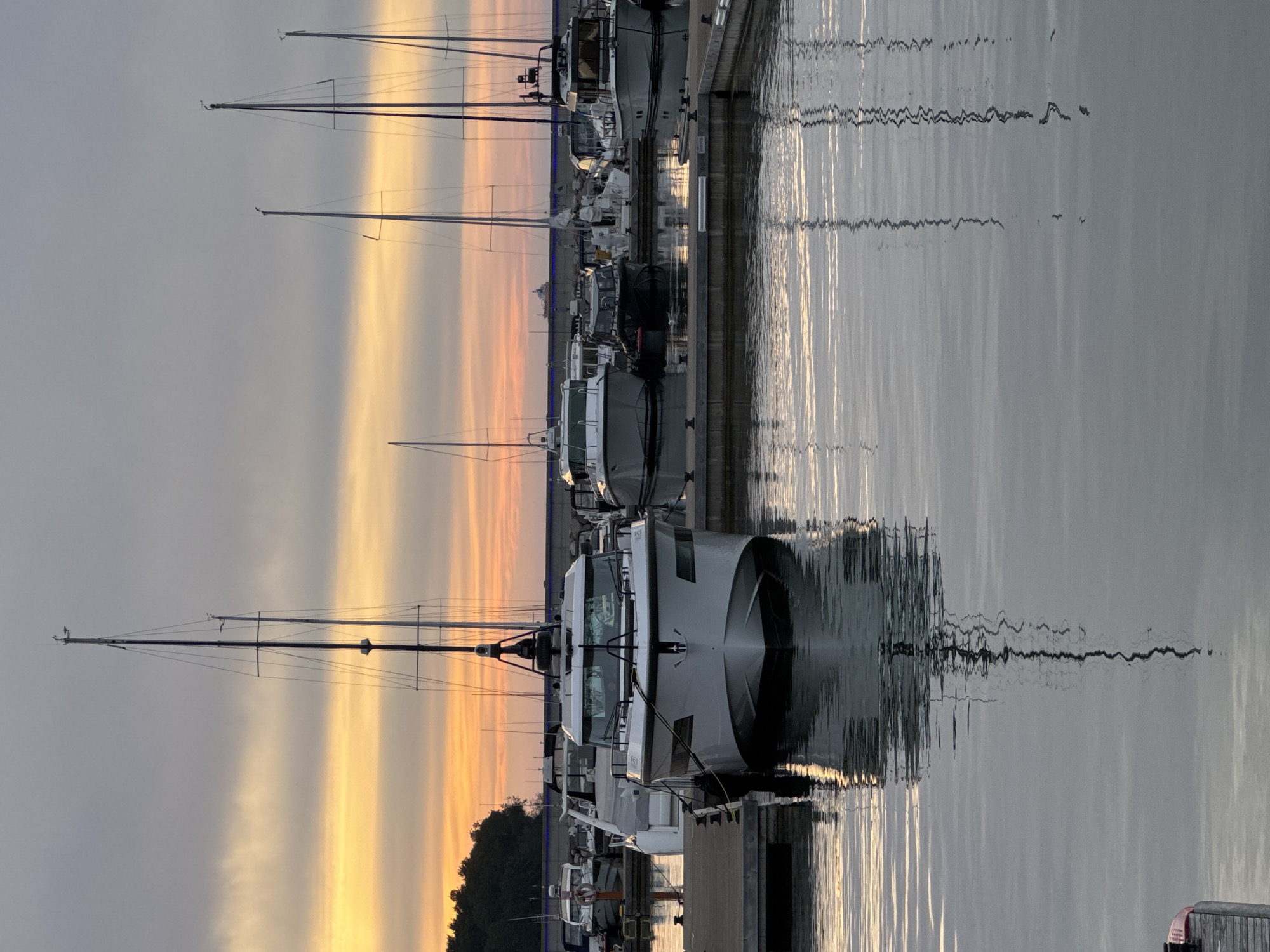 The image shows a marina in Tallinn, Estonia, at sunrise. The sky is filled with colorful clouds in shades of orange, yellow, and gray. Several boats are docked in the marina, including a large white motor yacht in the foreground. The water is calm and reflects the sky and the boats. In the background, there is a line of trees and a stone wall. A distant cargo ship can be seen on the horizon. The scene conveys a sense of tranquility and the beginning of a new day.