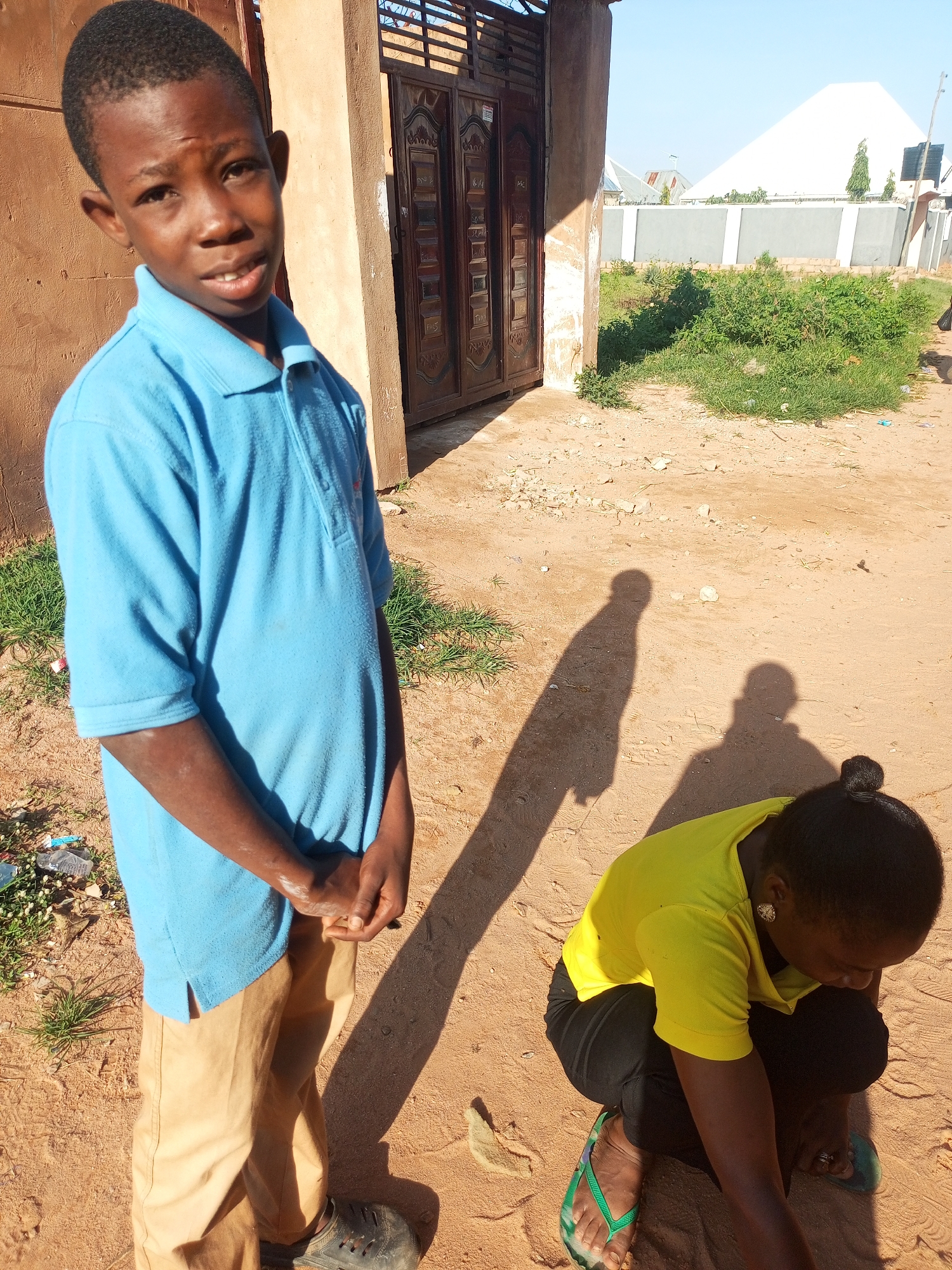 In Tempure, Nigeria, a young boy stands in front of a building with a large, ornate gate. He is wearing a blue polo shirt and khaki pants. To his right, a woman is crouching in the sand, wearing a yellow shirt and black pants. The environment appears to be outdoors, with sandy ground, some scattered vegetation, and a clear sky. Shadows indicate the presence of sunlight. In the background, more buildings and a wall are visible. The overall scene suggests a casual, everyday setting in a Nigerian city.