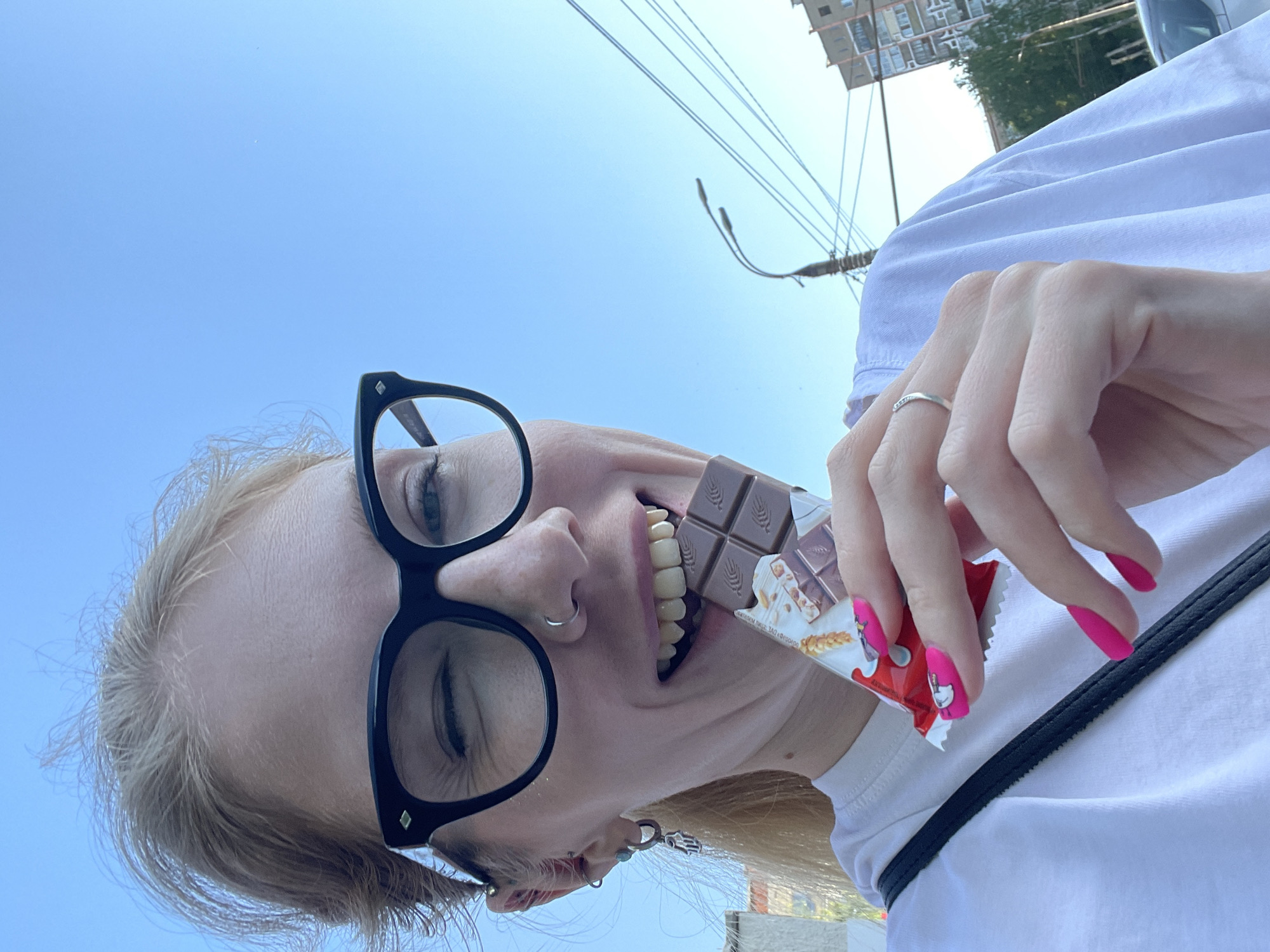 A fair-skinned person with blonde hair is holding a partially unwrapped chocolate bar in their mouth. They are wearing black-rimmed glasses and a white t-shirt with a black strap. Their nails are painted bright pink. The location appears to be outdoors in Izhevsk, Russia, based on the Cyrillic text on the chocolate wrapper. Buildings and overhead power lines are visible in the background under a clear blue sky. The photo is taken from a low angle, looking upwards.
