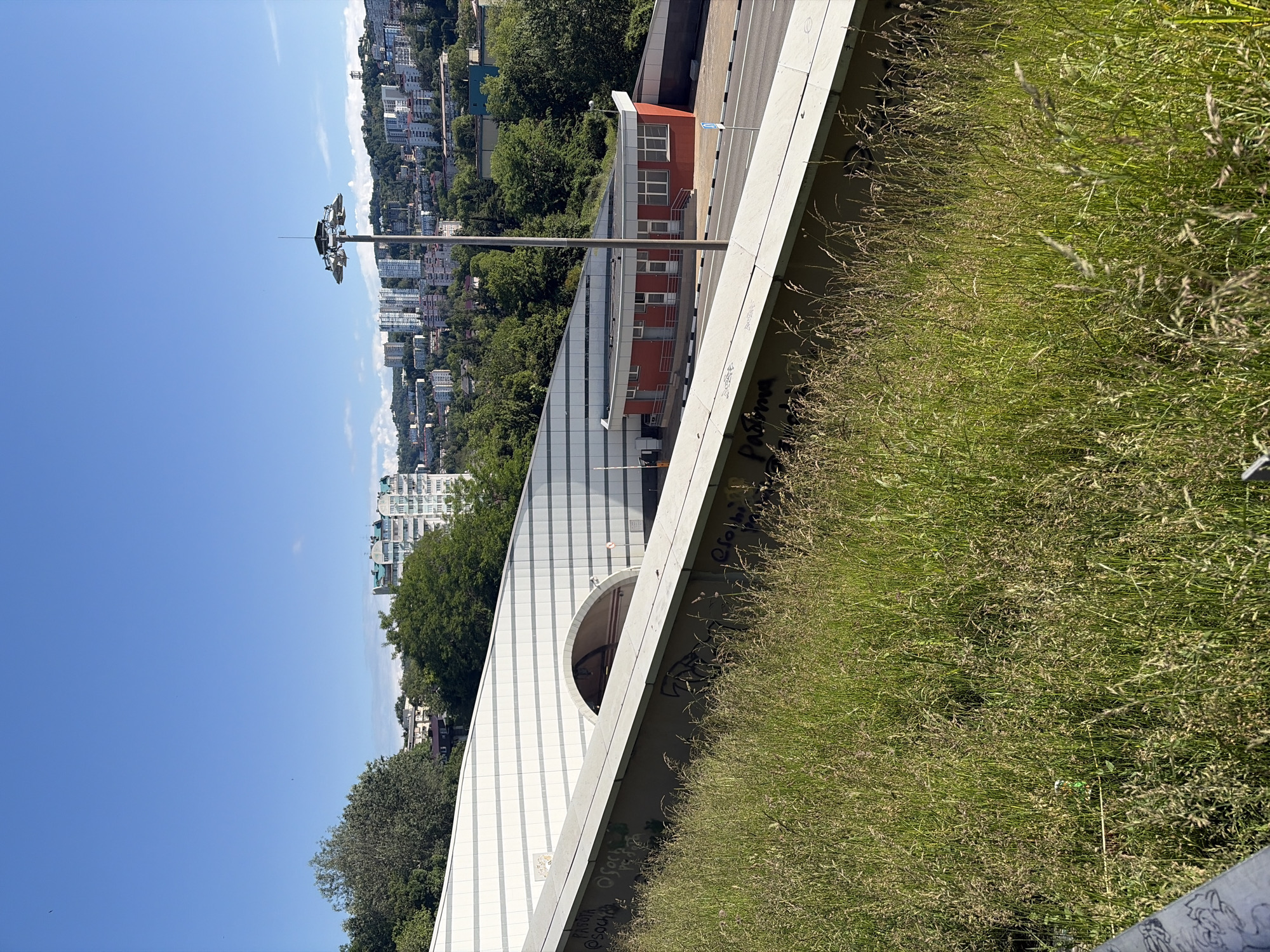 The image displays a daytime view of a landscape in Sochi, Russia, combining urban infrastructure with natural elements under a clear blue sky.
In the foreground, a verdant slope covered in tall, wild grass dominates the lower right portion of the frame. A concrete barrier or retaining wall runs horizontally along the edge of this grassy area, featuring visible graffiti. Some legible text includes "Pascha" and stylized markings that resemble "@soch" and possibly "OSOCHI."
The midground is occupied by a large, modern structure, likely part of a road or rail network, such as an overpass or tunnel entrance. This structure has a sloped, paneled surface in shades of grey and white, and features a distinctive arched opening. Above this structure, a paved road or service path is visible. Adjacent to the road stands a small, rectangular building with a reddish-brown exterior and white window frames, topped with a red roof, possibly serving as a utility building or control station. A tall metal pole equipped with several floodlights extends upwards from the road level.
The background reveals a panoramic view of a city sprawling across hills. Numerous multi-story buildings, appearing to be residential or commercial, are nestled among dense green trees and vegetation that cover the undulating terrain. The sky above is a brilliant, clear blue with only minimal, faint cloud formations, indicating a sunny and bright day. No people or specific activities are visible, creating a static yet expansive depiction of the city's interface with its natural surroundings.