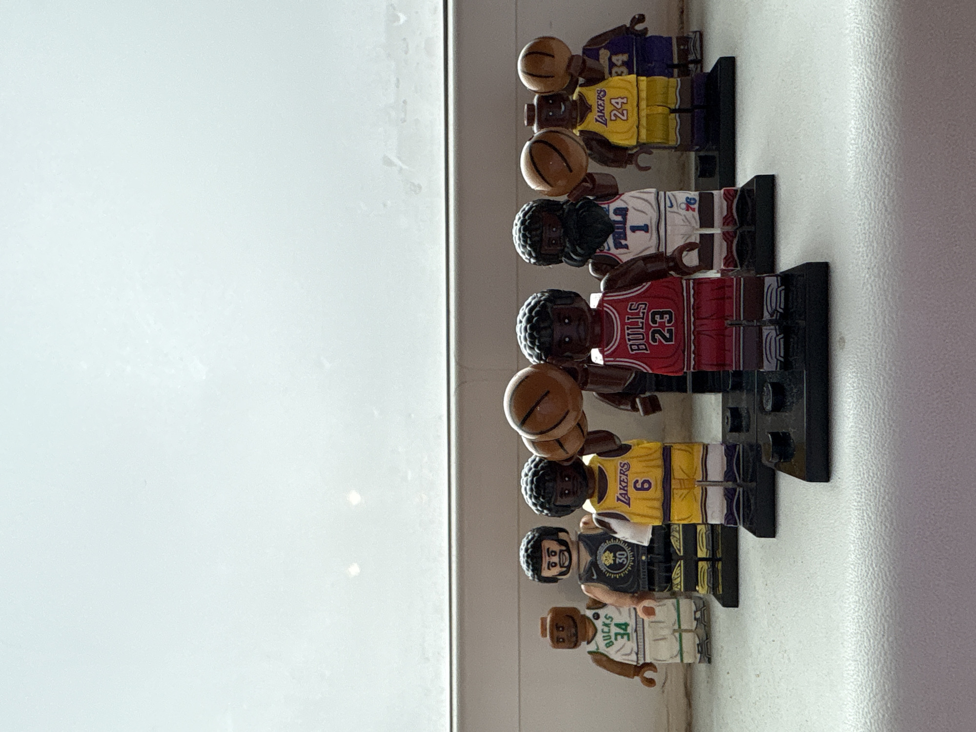 The image shows a collection of Lego basketball player figurines arranged in a row. The figurines are wearing jerseys representing different teams and players, including the Los Angeles Lakers (numbers 24 and 6), Philadelphia 76ers (number 1), Chicago Bulls (number 23), Golden State Warriors (number 30), and Milwaukee Bucks (number 34). Some figures are holding a basketball. The Lego figurines are positioned near a white surface.