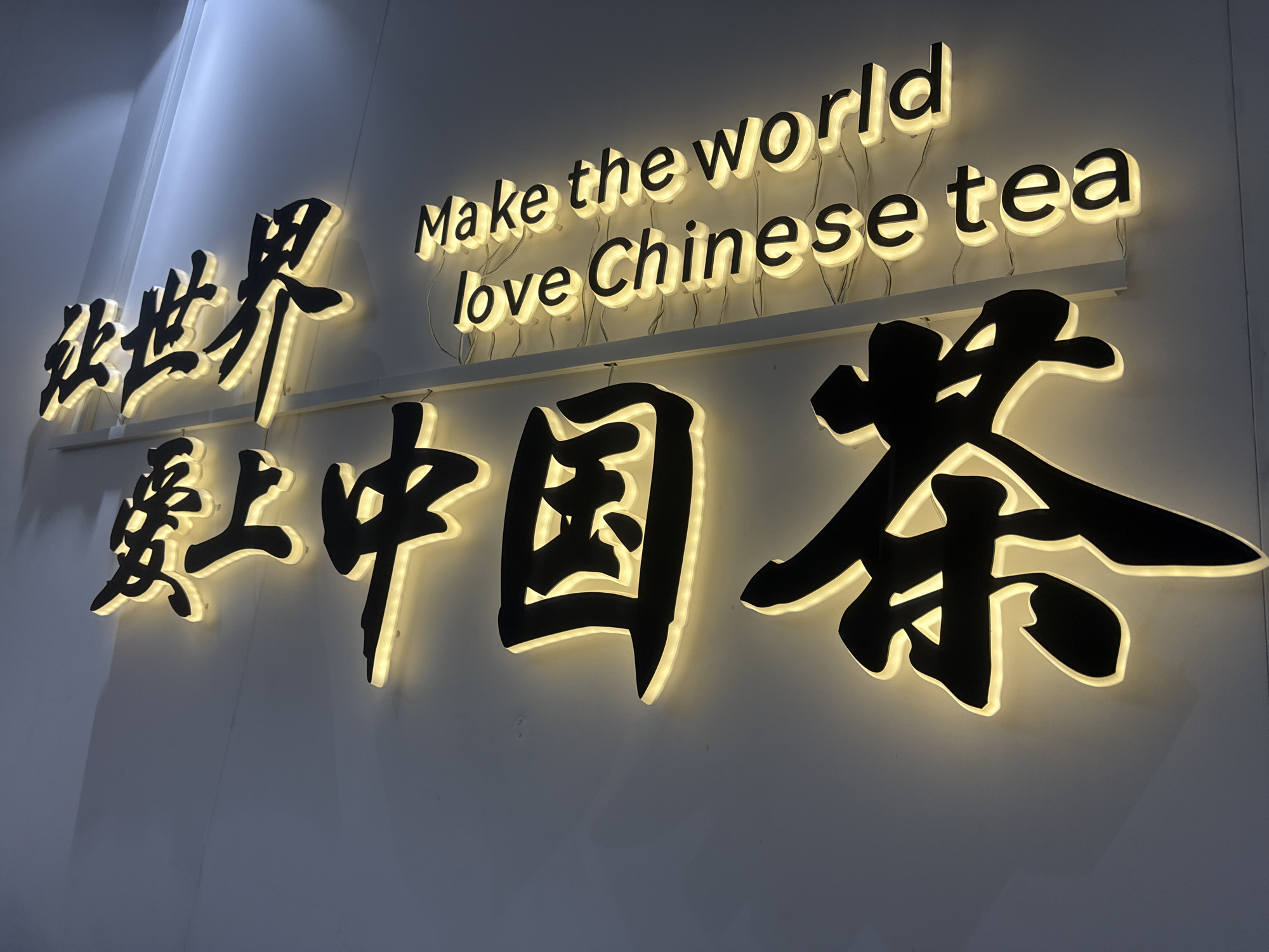 An illuminated sign, featuring both English and Chinese text, is mounted on a light-colored wall, likely indoors. The lighting in the scene is dim, causing the backlit sign to stand out prominently.
The upper part of the sign displays the English text: "Make the world love Chinese tea." These words are formed by individual black block letters, each surrounded by a soft, white-yellow halo of light emanating from behind, creating a glowing effect against the wall. Some thin electrical wires connecting these letters are visible.
Below and to the left of the English text, the same message is presented in large, stylized Chinese characters: "让世界爱上中国茶" (Ràng shìjiè ài shàng Zhōngguó chá). These characters are rendered in a calligraphic style, appearing as black shapes with a similar warm, soft white backlighting that provides a striking contrast with the dim background. The larger size and artistic style of the Chinese characters give them a more emphatic visual presence.
The sign promotes Chinese tea with a global message. Given the location context of Yekaterinburg, Russia, the sign serves as a cross-cultural advertisement or declaration, aiming to introduce or popularize Chinese tea to an international audience. The warm glow of the sign creates an inviting and somewhat elegant atmosphere.