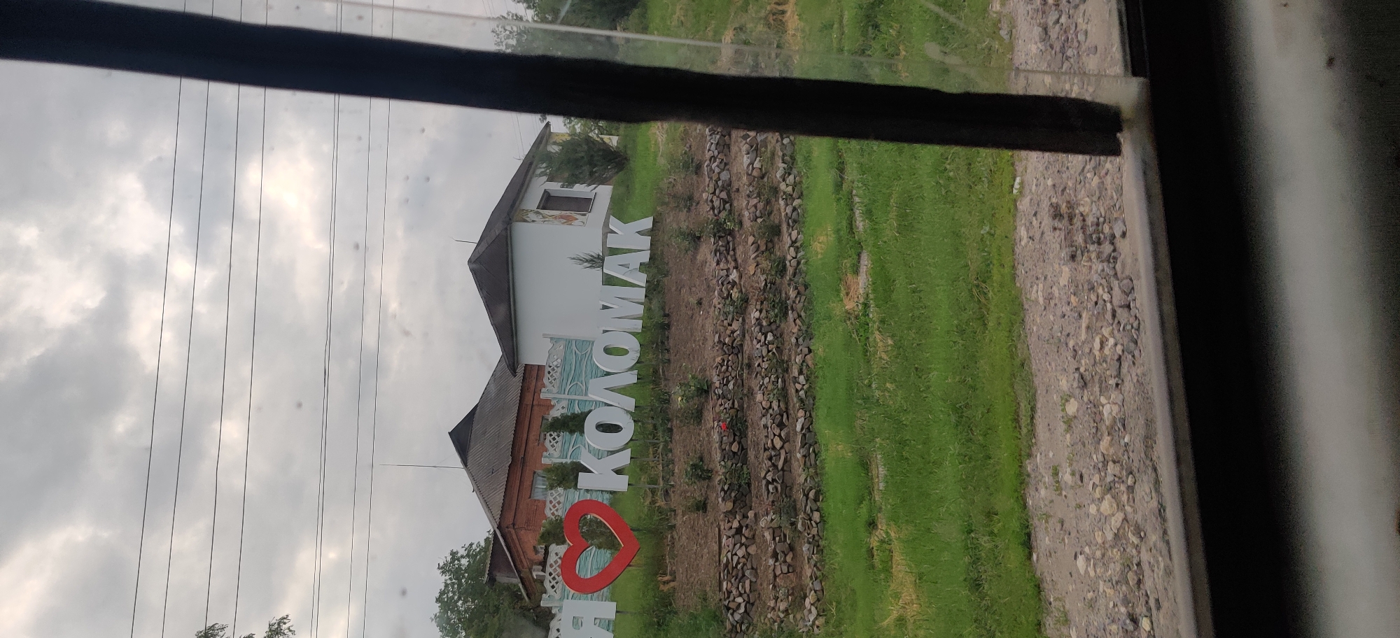 The image displays an outdoor scene, likely captured from a vehicle given the dark frame and visible smudges/reflections on what appears to be a window. The main focal point is a large, white monument sign set on the ground, prominently featuring the text "Я ❤️ КОЛОМАК" in Ukrainian Cyrillic. This translates to "I love Kolomak," with a vibrant red heart symbol replacing the word "love."
Behind the sign, two buildings are visible. The left building appears to be an older structure, possibly made of brick, with a dark, sloped roof. To its right, a light-colored, presumably white, building also has a dark, sloped roof and features decorative murals or paintings on its exterior walls, including a stylized window and a blue, wave-like pattern near its base.
The ground surrounding the sign and buildings consists of patches of green grass interspersed with areas of loose stones or rocks. The sky above is heavily overcast, suggesting a cloudy daytime. Several dark, thin power lines or utility wires traverse the upper part of the image against the gray sky. The scene is static, with no people or discernible activities taking place. The presence of the "I love Kolomak" sign acts as a clear location cue for Kolomak, Ukraine, conveying a sense of local pride.