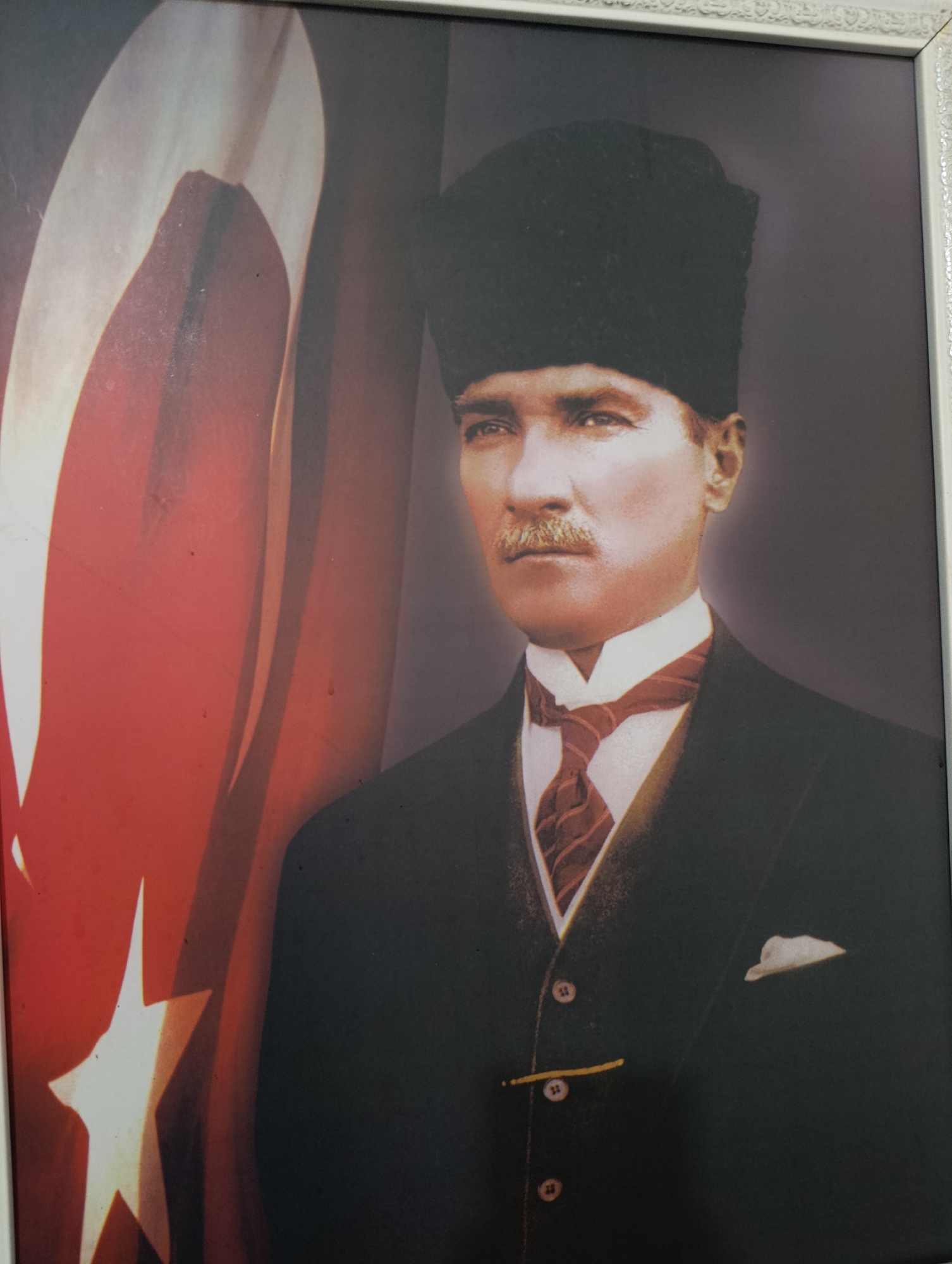 A framed portrait depicts a man in a dark suit and a black, fur-brimmed hat, standing in front of a Turkish flag. The man has a mustache and is wearing a white collared shirt with a red and brown striped tie. A white pocket square is visible in the breast pocket of his suit jacket. The flag, which is partially obscured by the man, features a red background with a white star and crescent moon. The image is illuminated, and the framing is ornate. The overall scene is a formal portrait.