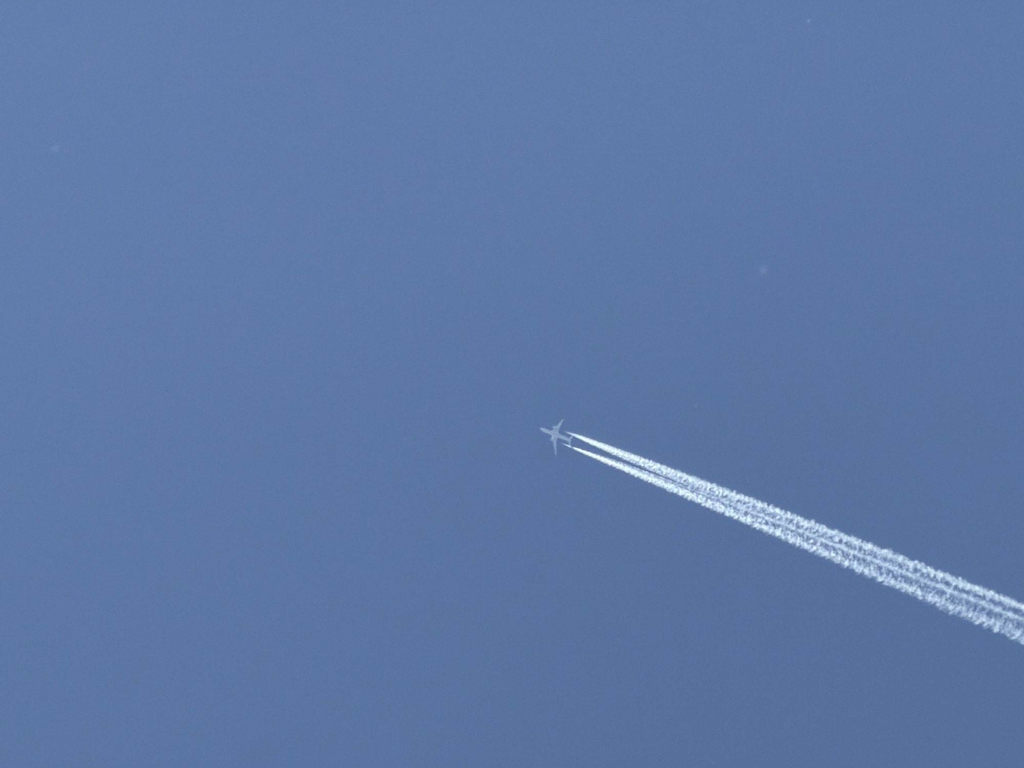 The image shows a clear, blue sky. An airplane is visible in the center-right portion of the image, leaving two distinct contrails behind it. The plane is angled upwards and to the right. The location is unspecified and cannot be identified as Izhevsk, Russia, or any other location.
