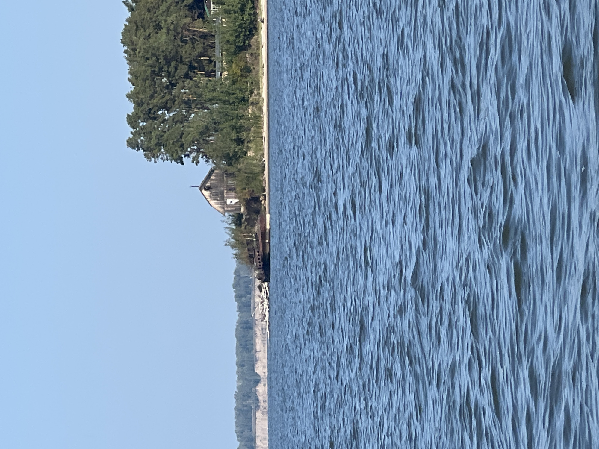 An eye-level, full shot captures a wide expanse of rippling blue water in the foreground. The middle ground reveals a sandy shoreline dotted with trees and vegetation. A weathered, wooden building with a steep roof sits on the embankment, partially obscured by foliage. To the left of the building, a rusty, partially submerged shipwreck is visible, with debris scattered around it. In the background, a distant cliff face with visible erosion rises, topped by a line of trees. The sky is clear and pale blue. No people are clearly visible. The scene suggests a remote, possibly abandoned coastal area. The provided location information indicates Borovoye, Russia.