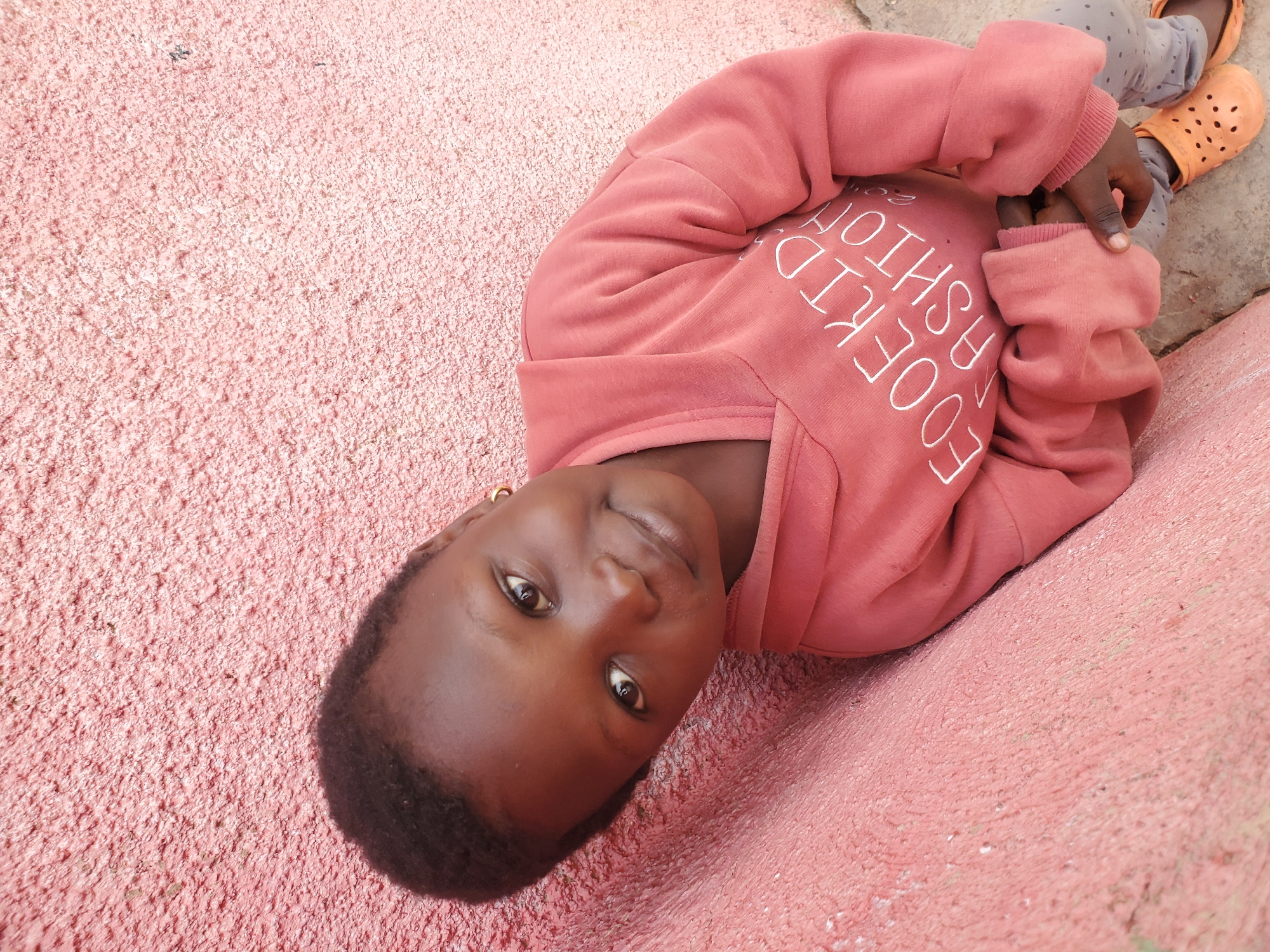 A young child is leaning against a textured pink wall, looking towards the camera with a slight smile. They are wearing a coral-colored hooded sweatshirt with white lettering that reads "EOOEKID FASHION 2022." The child also has on grey patterned leggings and orange perforated clogs. The background is the pink wall and a grey ground surface. The image appears to have been taken outdoors in a casual setting, likely a residential area in the city of Jos, Nigeria, given the characteristic rough pink plaster common in some Nigerian buildings and the overall ambiance.