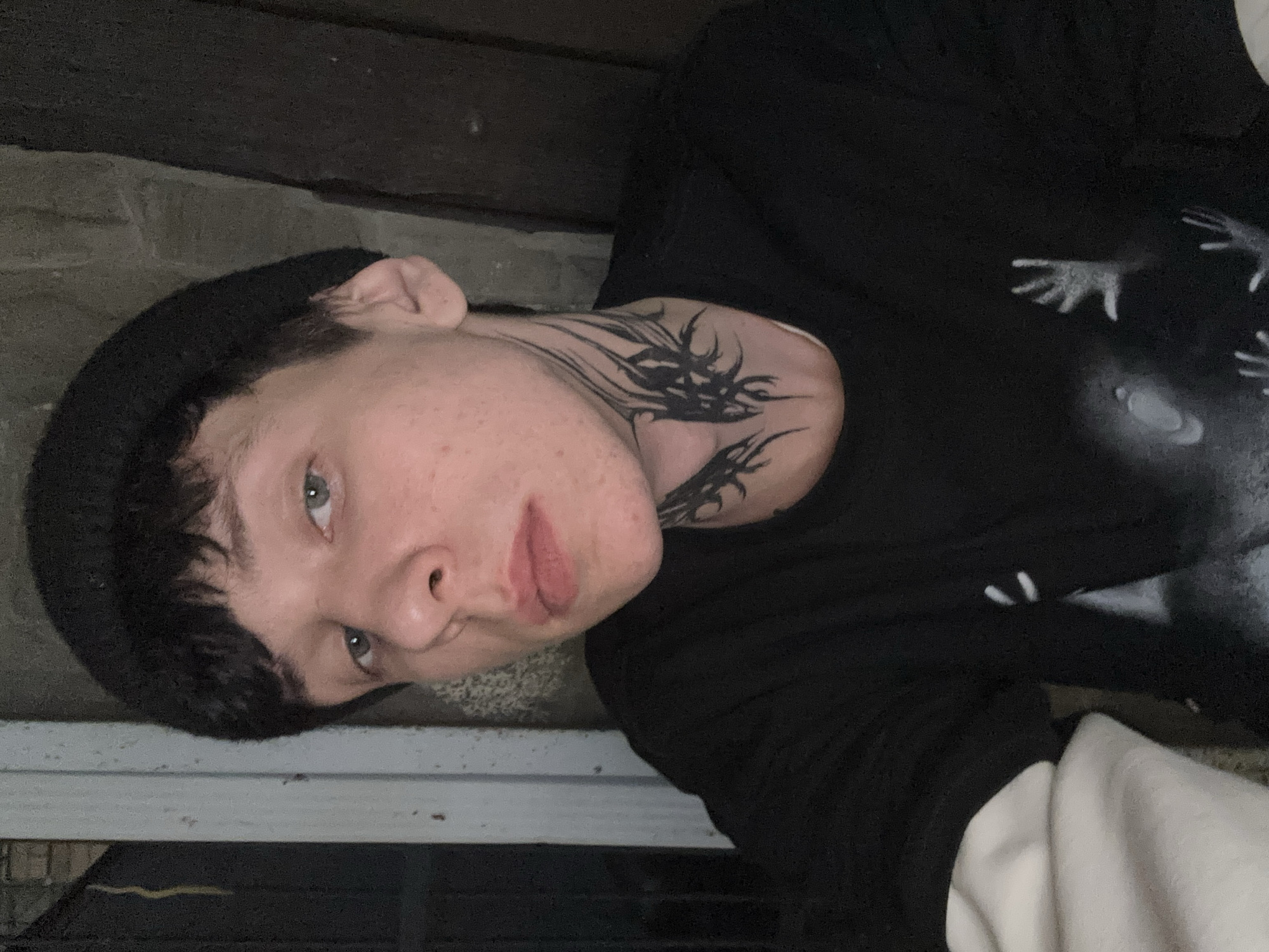 A young, fair-skinned person with short, dark hair and a black beanie is positioned in the frame, displaying a dark, abstract neck tattoo. The person's eyes are light-colored. They are wearing a dark-colored shirt with a graphic design of hands and a celestial body in white or light gray, with the hint of a lighter-colored undershirt visible at the bottom. The individual appears to be indoors, next to a light-colored ledge or surface. The image's orientation suggests it may have been taken from a low angle and rotated. The specific location within Saratov, Russia is not discernible from the image.