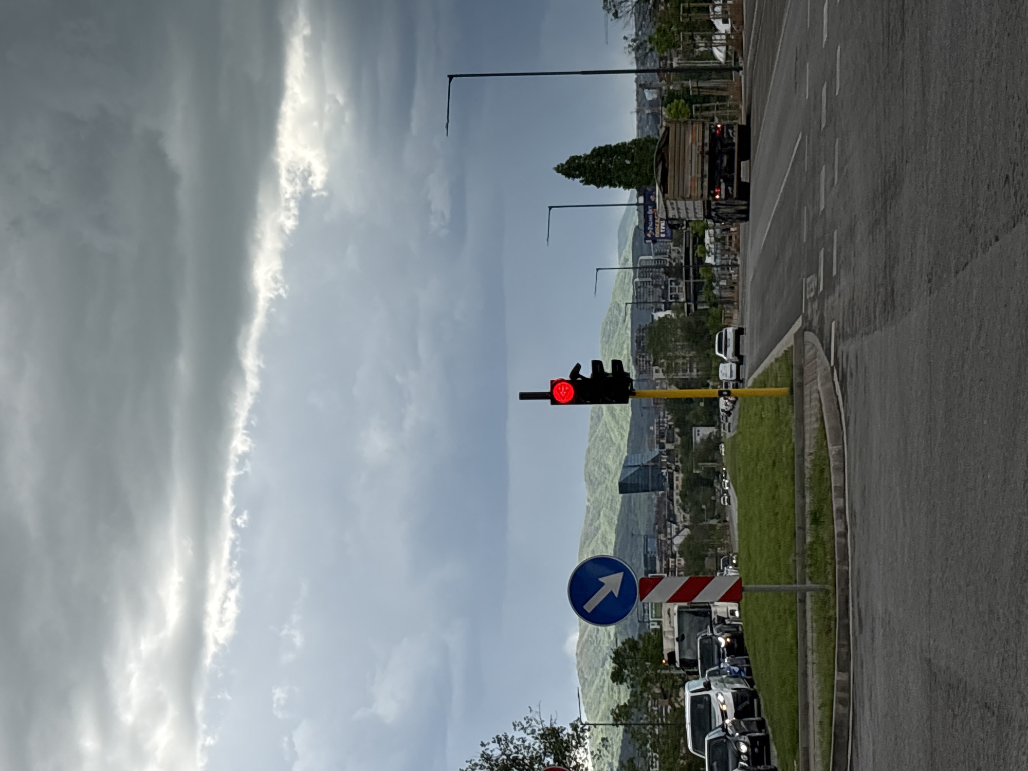 The image depicts a street scene in Sofia, Bulgaria. A red traffic light is visible, indicating stopped traffic. A blue circular sign with a white arrow pointing to the left indicates the direction of the road. The road is paved with asphalt. In the background, there are buildings and mountains. The sky is cloudy. A line of cars is waiting at the traffic light. A truck is further ahead on the road. Street lights are visible along the roadside. There is a grassy area alongside the road.