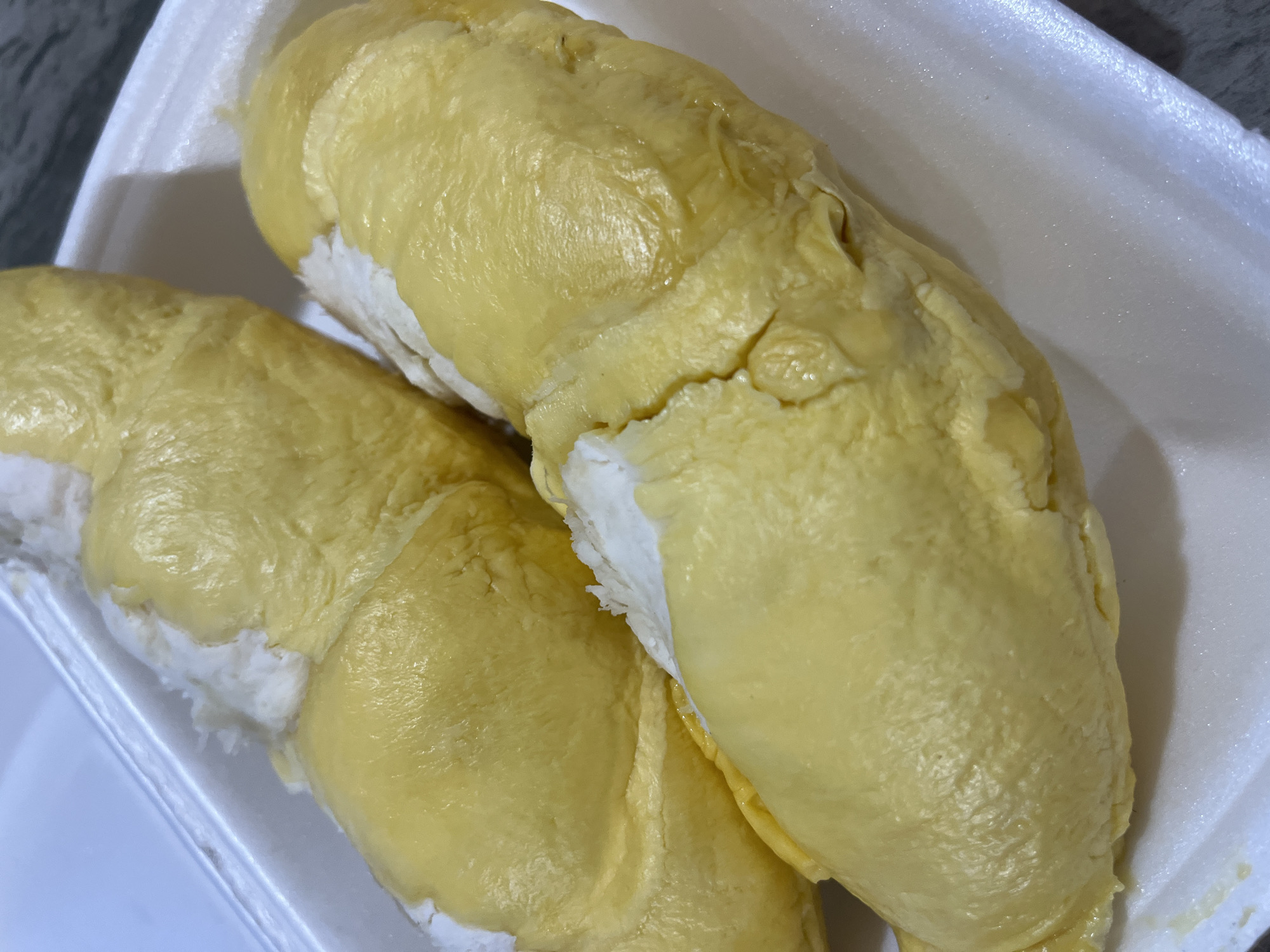 The image shows two pieces of durian fruit in a white styrofoam container. The durian flesh is a pale yellow color with a slightly wrinkled texture. Some white, fibrous material is visible at the base of each piece. The container is sitting on a dark, possibly granite surface. The image appears to be taken indoors with natural lighting. The location in Bac Nha Trang Ward, Vietnam is implied, not directly visible in the image.